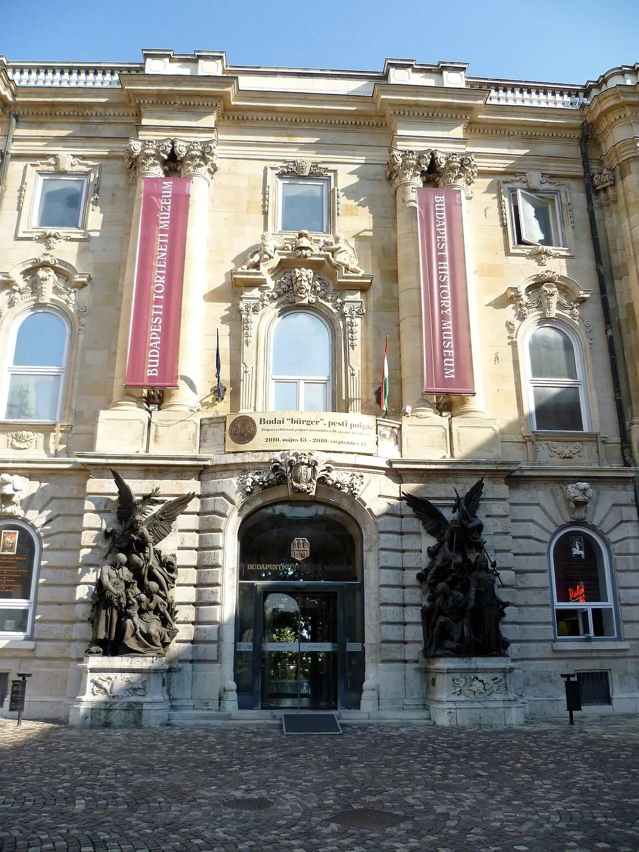 The main entrance of the Budapest History Museum on the Lion Courtyard, with banners reading Budapesti Történeti Múzeum and Budapest History Museum either side of the arched doorway