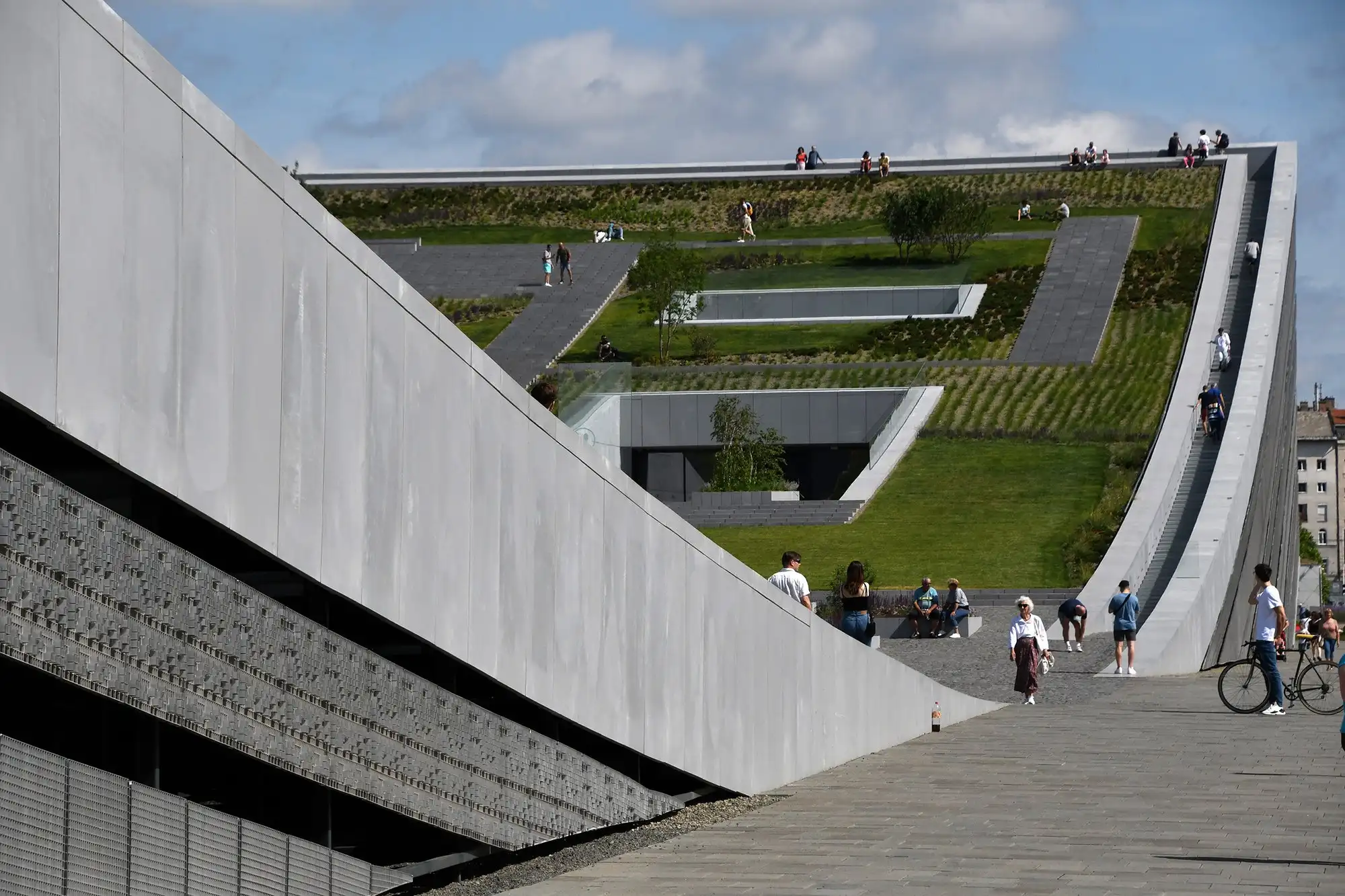 Visitors on the planted roof garden of the Museum of Ethnography in Budapest on a sunny day, walking up the steps along its curved edges, sitting at its foot and standing along the top