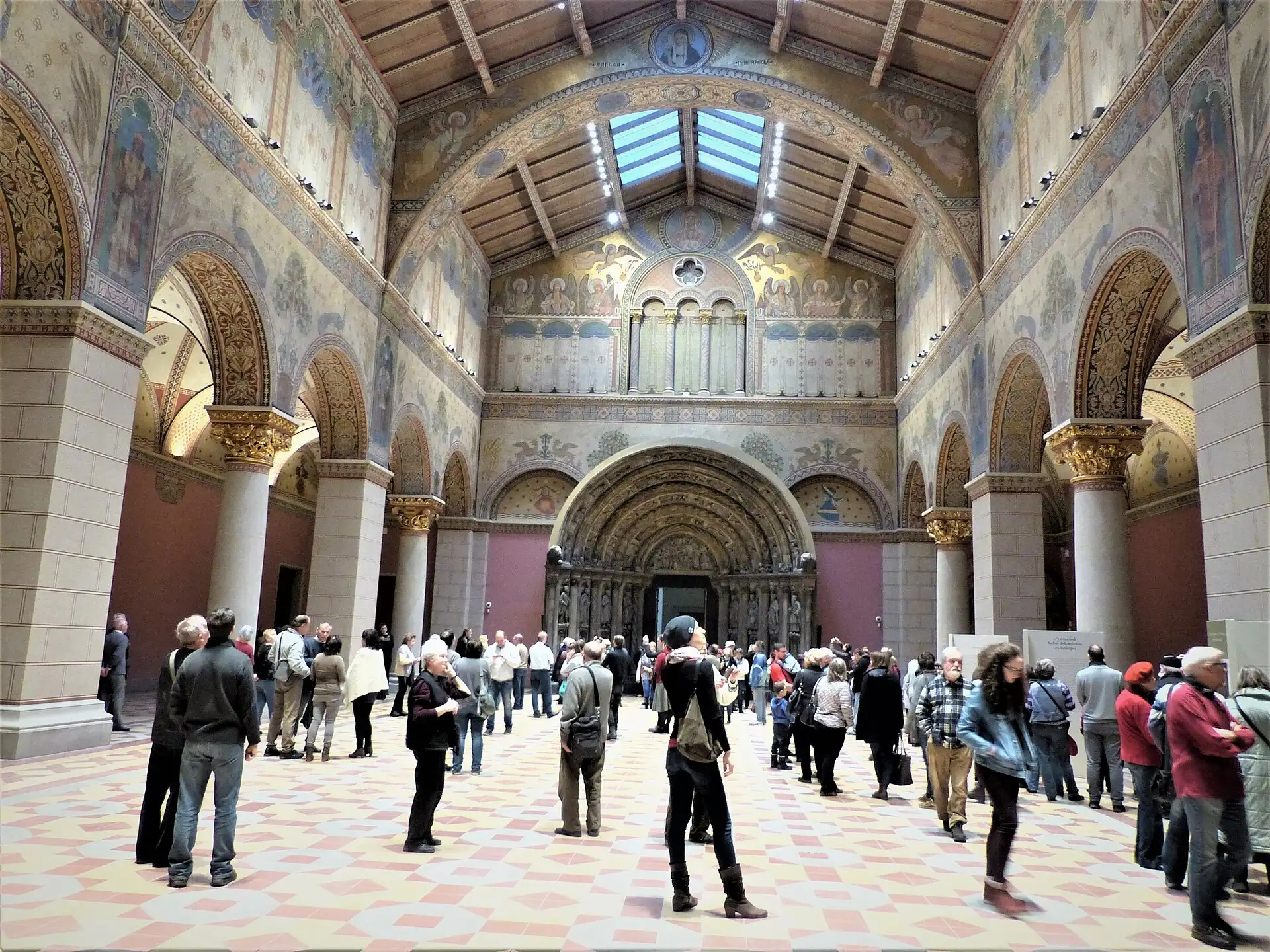 The Romanesque Hall of the Museum of Fine Arts in Budapest, a tall basilica-like room with painted arcading down both sides and visitors standing under the stone portal at the far end