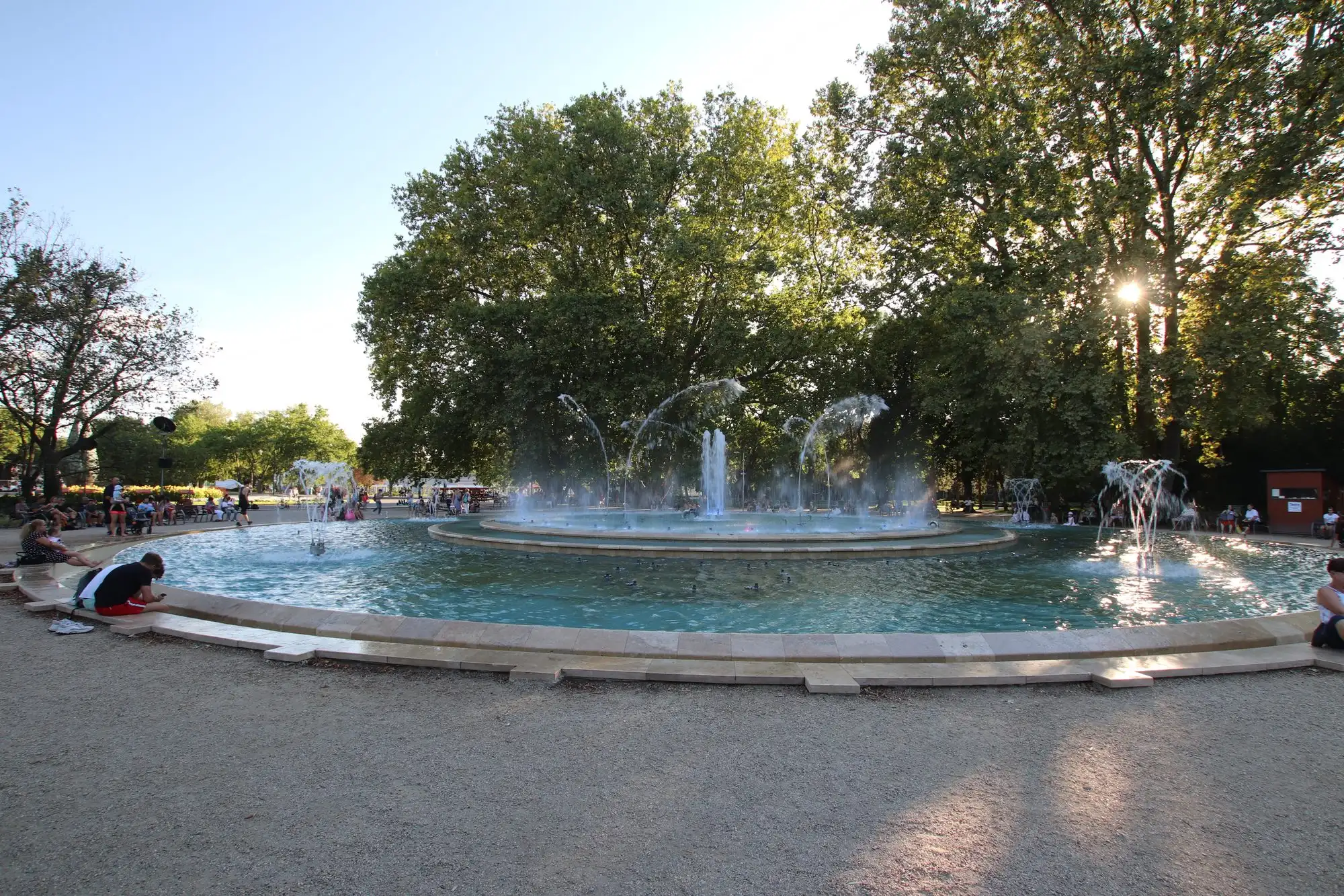 The musical fountain on Margaret Island in full flow, jets playing over the round basin with plane trees behind and people sitting on the stone rim