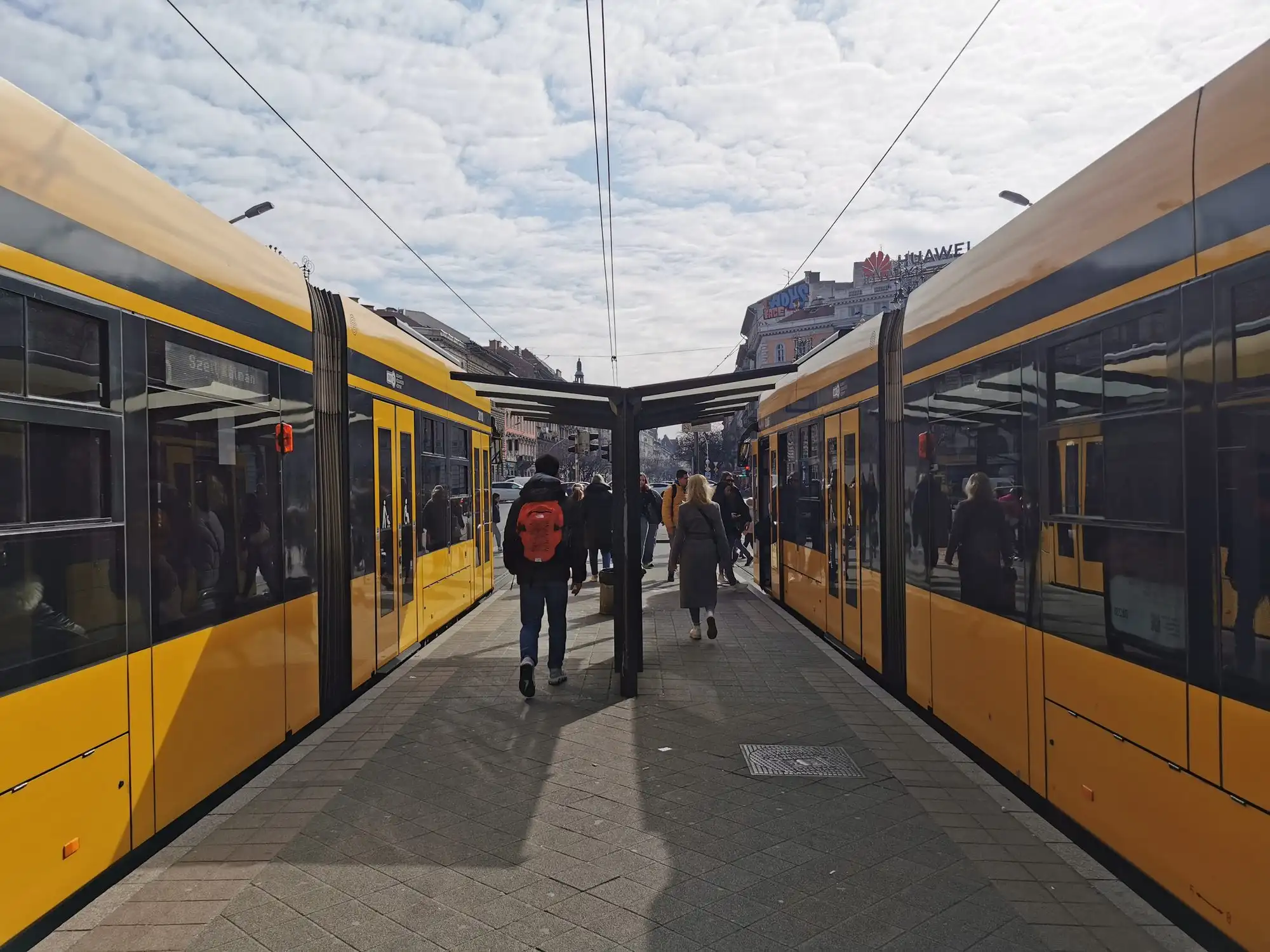 Two yellow trams at the island platform of the Oktogon stop on Budapest's Grand Boulevard, with passengers walking between them