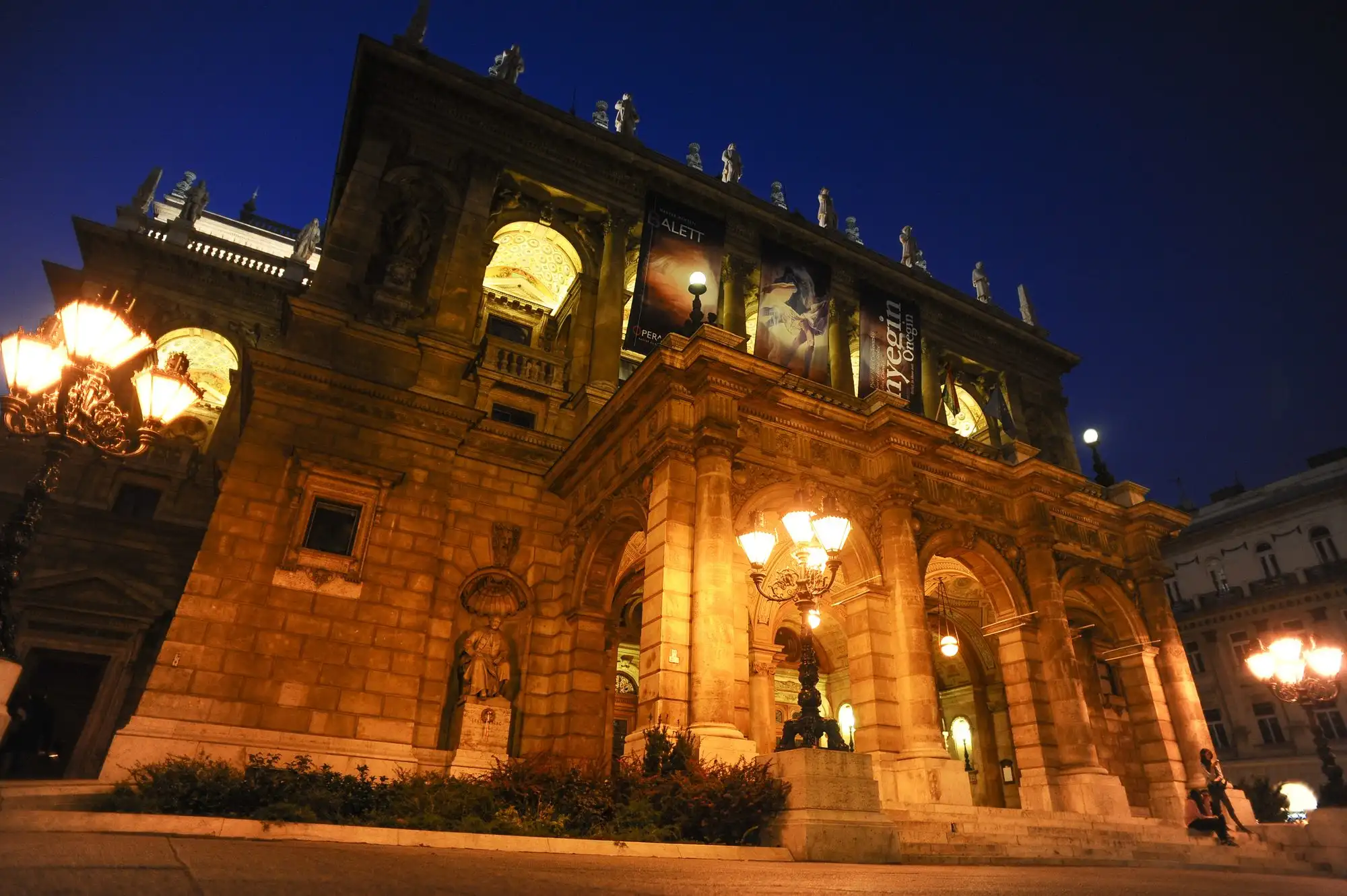The front of the Hungarian State Opera House lit up at night, with lamps glowing under the arcade and statues along the roofline