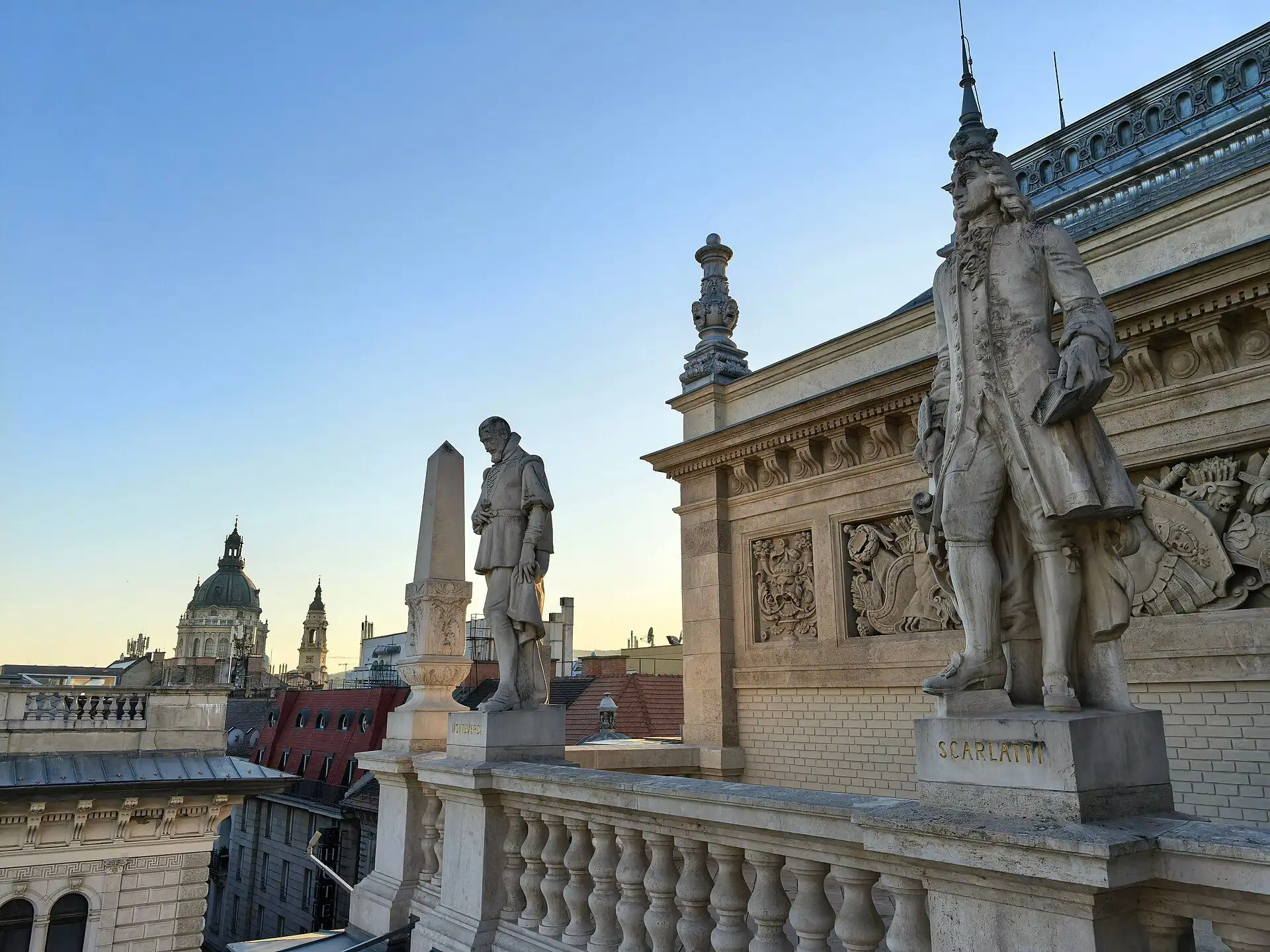 Composer statues standing along the parapet of the Hungarian State Opera House at dusk, with the dome of St Stephen's Basilica behind them