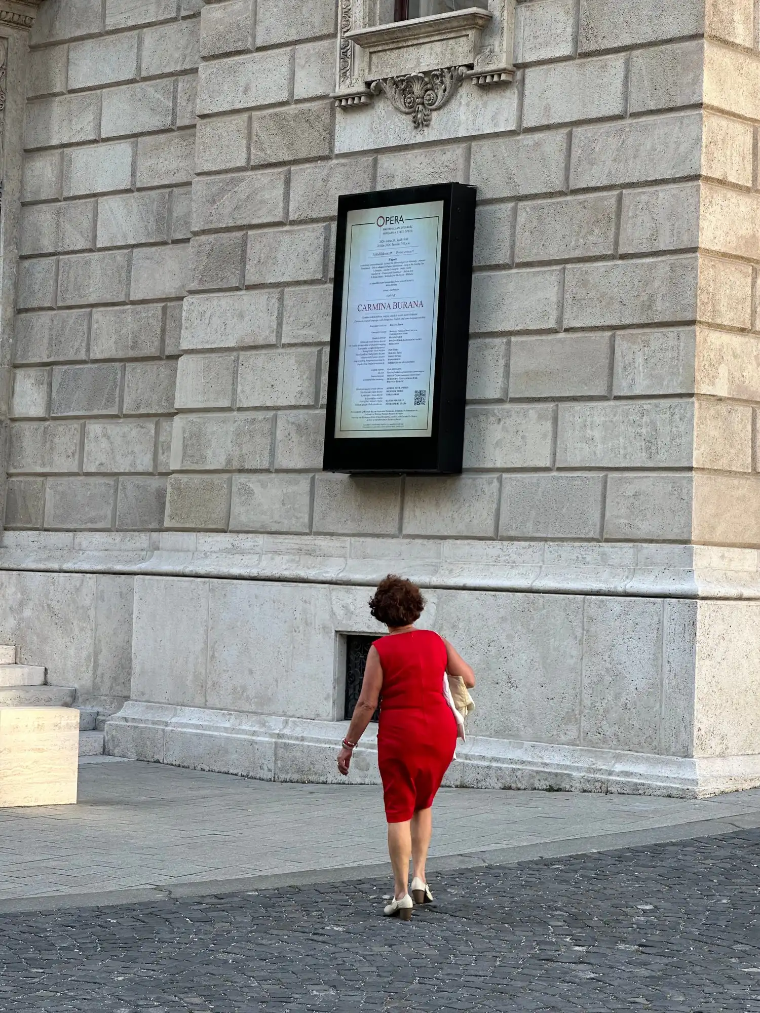 A woman in a red dress walking up to the evening's programme board on the stone wall of the Opera House on Andrássy út