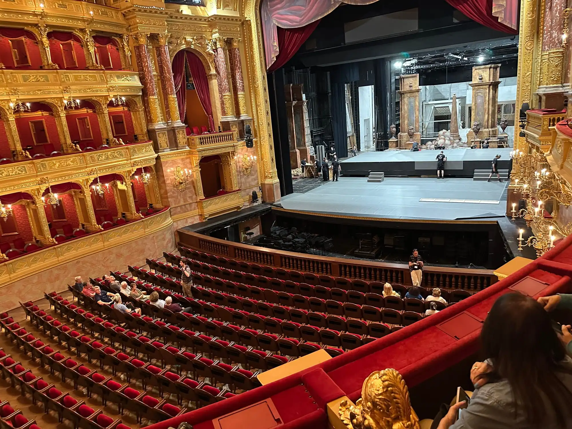 Stage crew setting up a production on the Opera House stage while tour groups sit in the stalls and in a box of the gilded auditorium