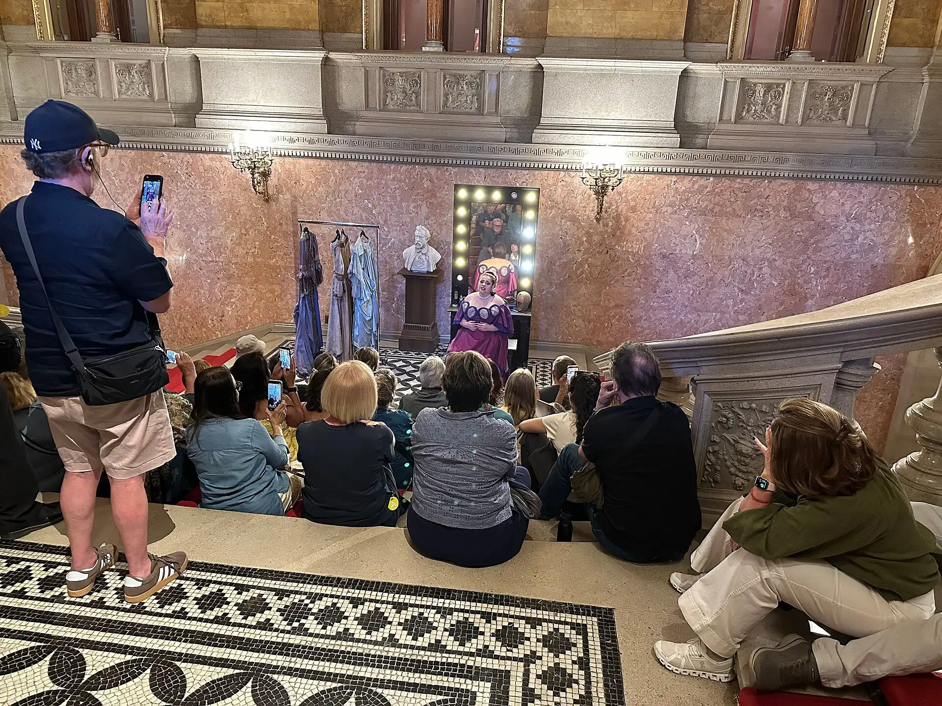A tour group sitting on the steps of the grand staircase watching two costumed opera singers perform on the landing