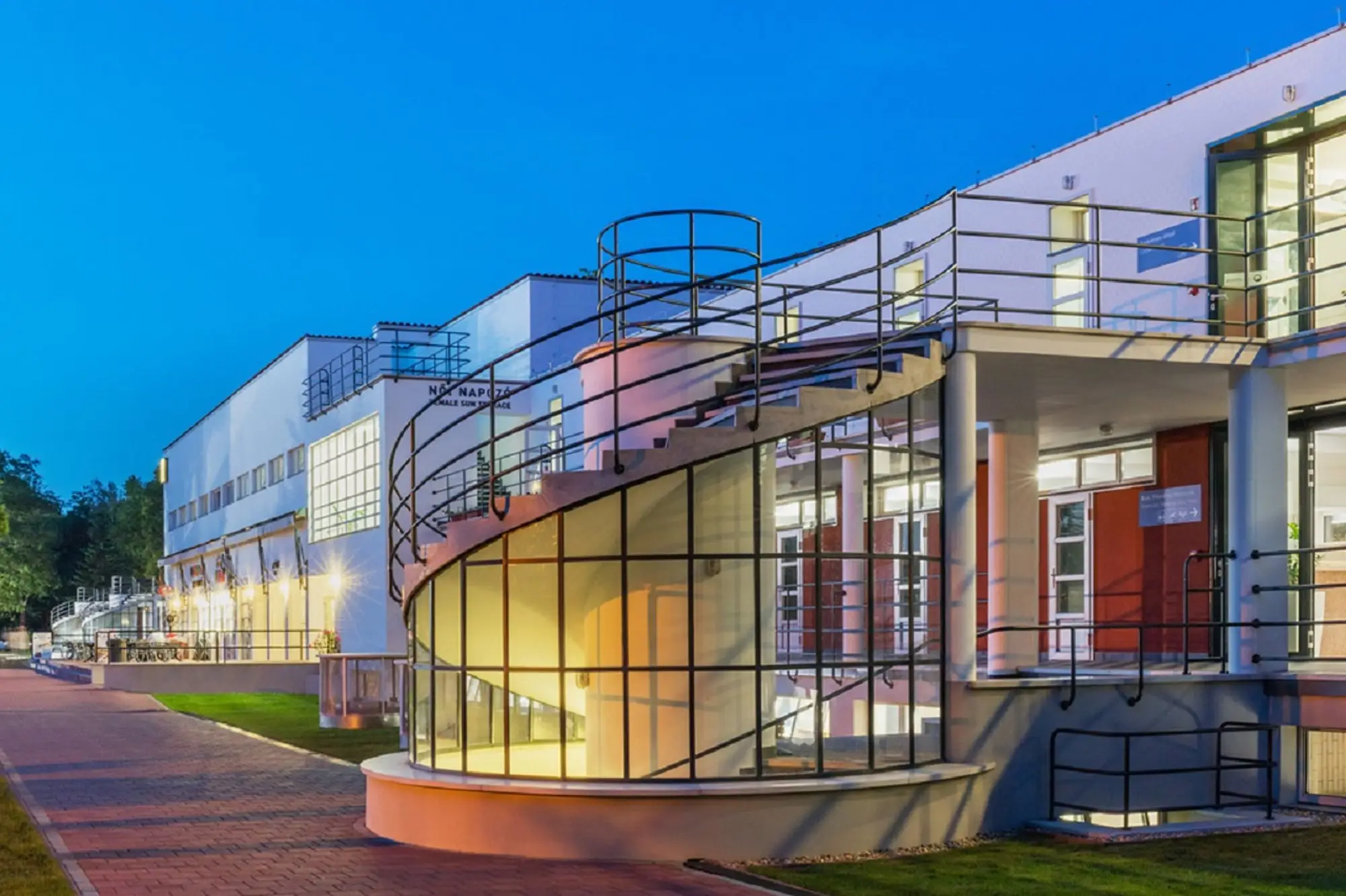 The white Bauhaus building of the Palatinus bath on Margaret Island at dusk, with its glass spiral staircase lit from inside