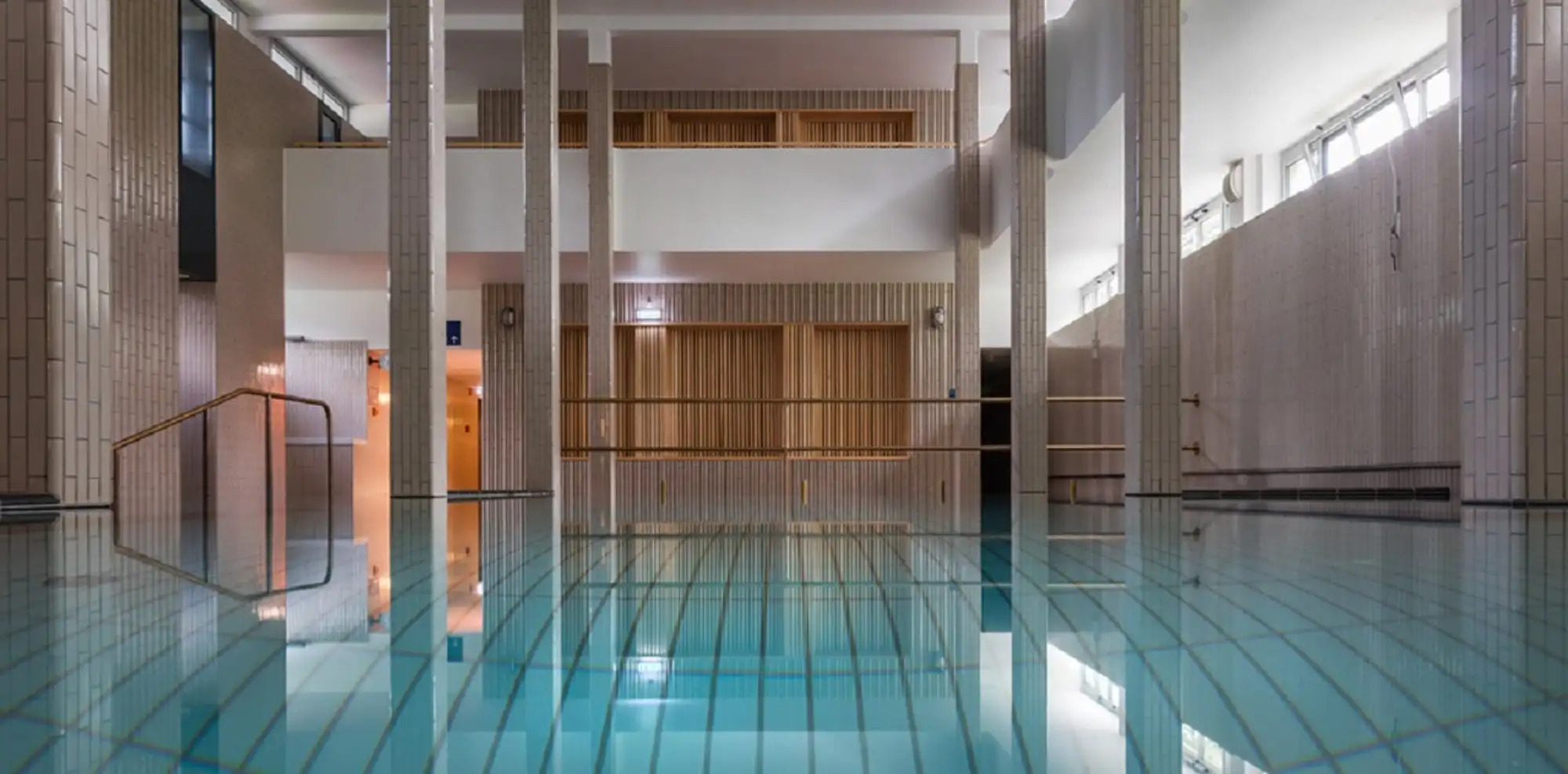 The indoor pool hall at Palatinus, still turquoise water between tall tiled columns, with a brass handrail at the steps