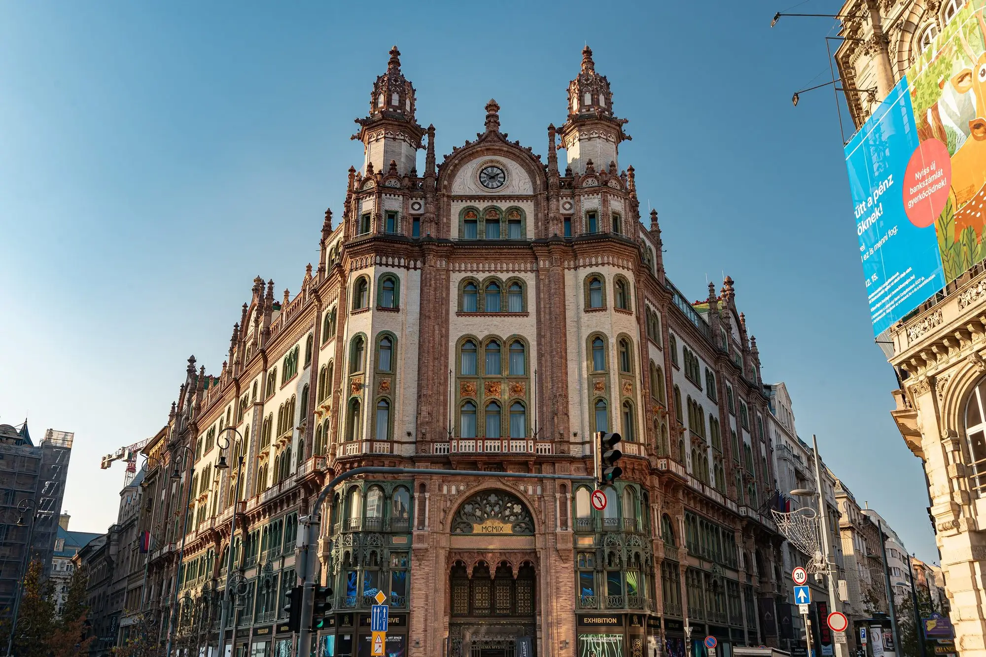 The Párisi Udvar building on Ferenciek tere in Budapest, its brick and stone facade and twin turrets seen from the square, with exhibition signs at street level