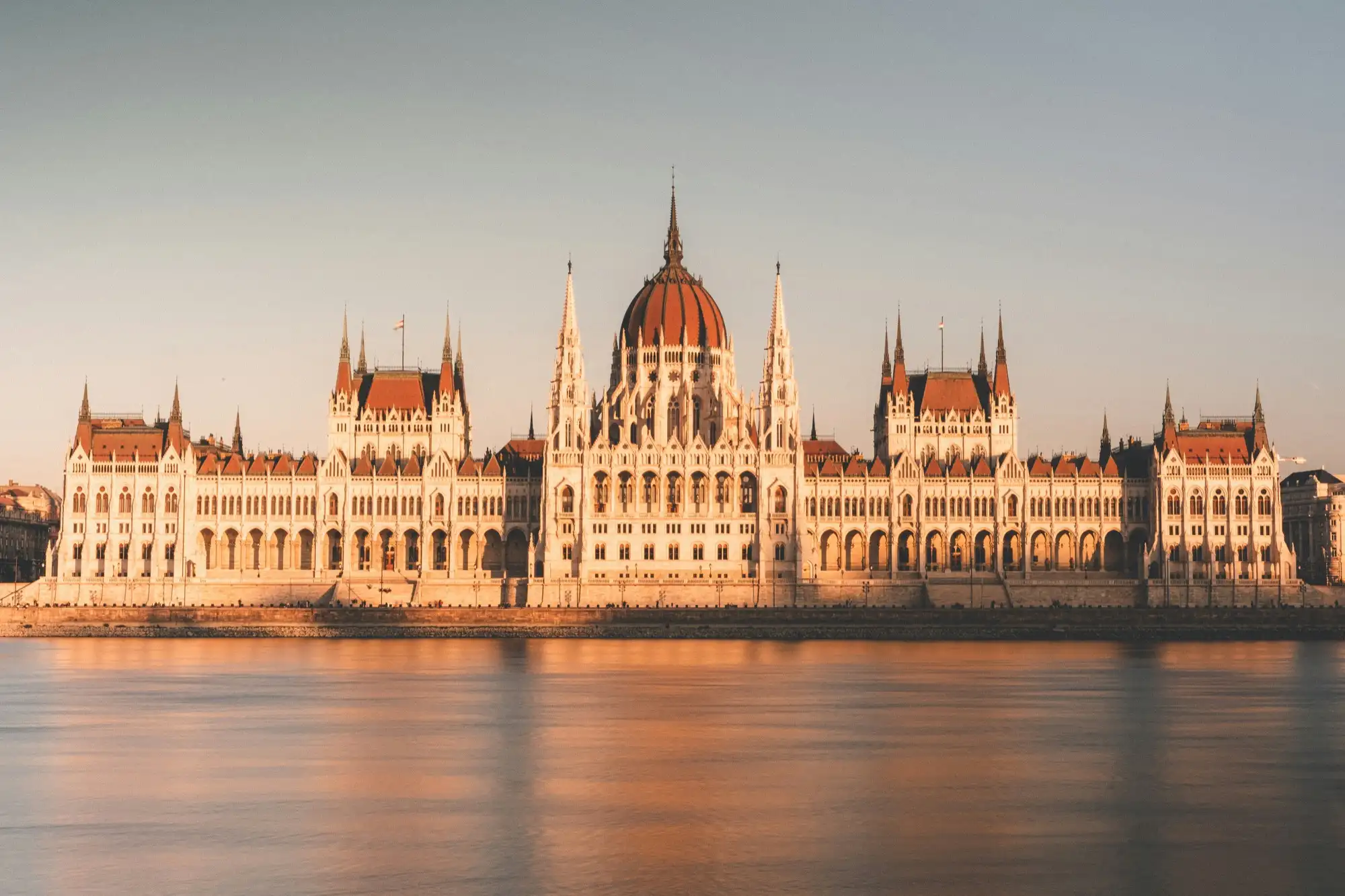 The Hungarian Parliament seen straight on across the Danube from the Buda bank, its facade lit by low golden sunlight above the calm river