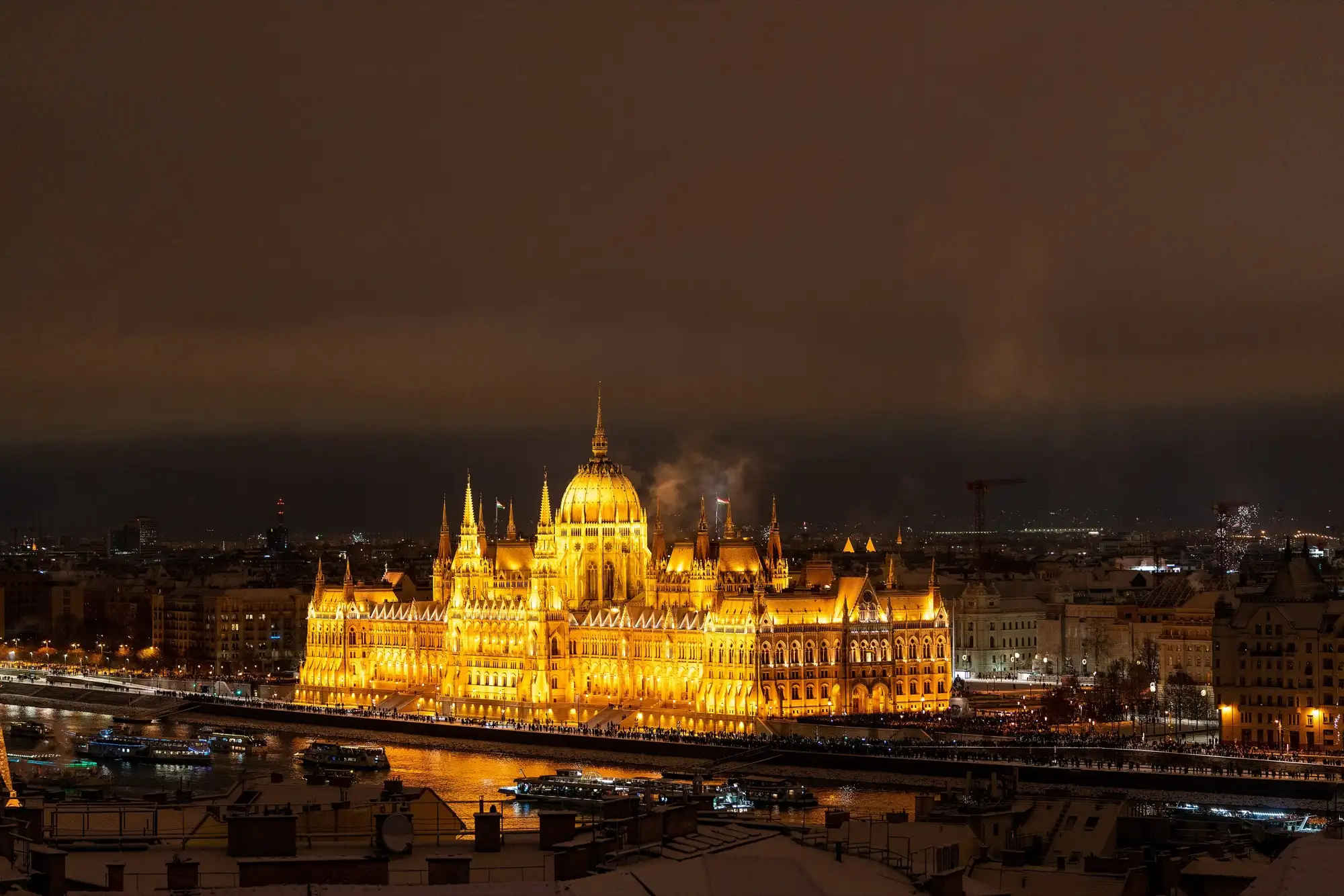 The Hungarian Parliament lit up gold across the Danube on a winter night, with snow on the rooftops in the foreground