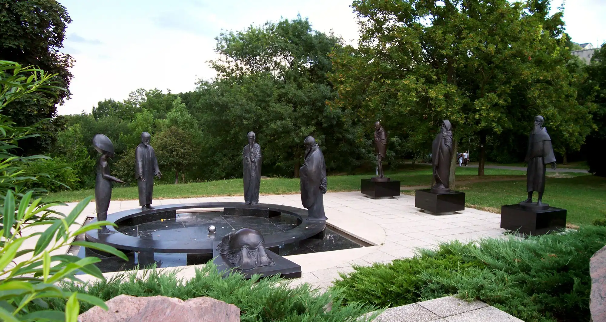 The Philosophers' Garden on Gellért Hill, a ring of bronze figures standing round a dark circular pool on a paved terrace, with low shrubs in front and trees behind
