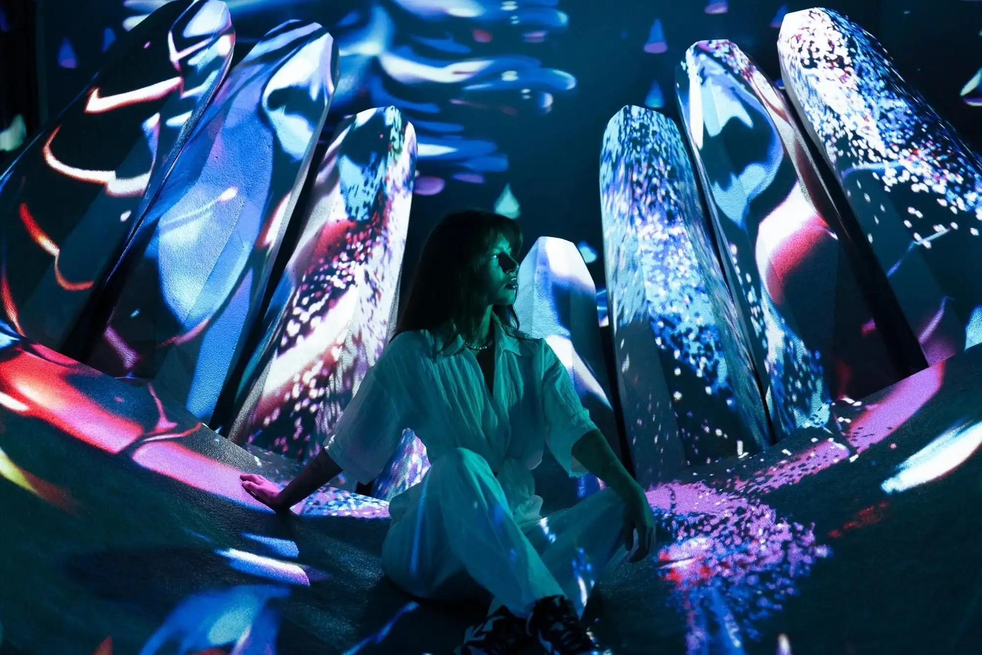 A visitor sitting among tall, angled white forms lit by blue and pink projections in a room at Cinema Mystica in Budapest