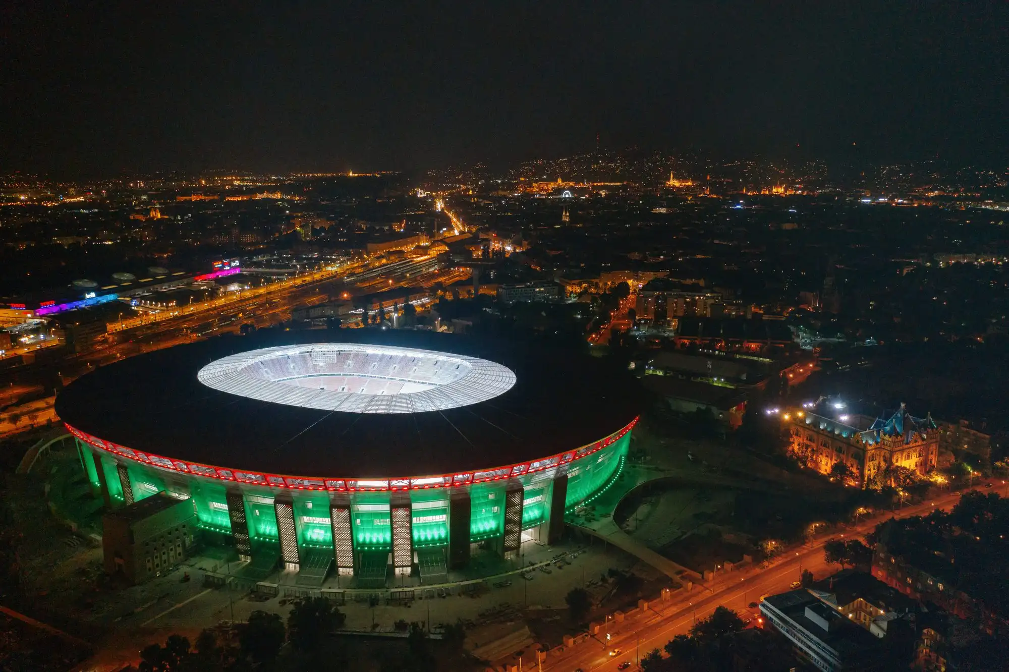 Puskás Aréna from the air at night, its facade lit in red, white and green, with the roads and the park around it