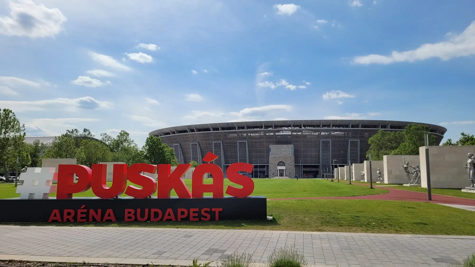 The red PUSKÁS ARÉNA BUDAPEST sign on the lawn of the Puskás Aréna Park, with the grey stadium behind it
