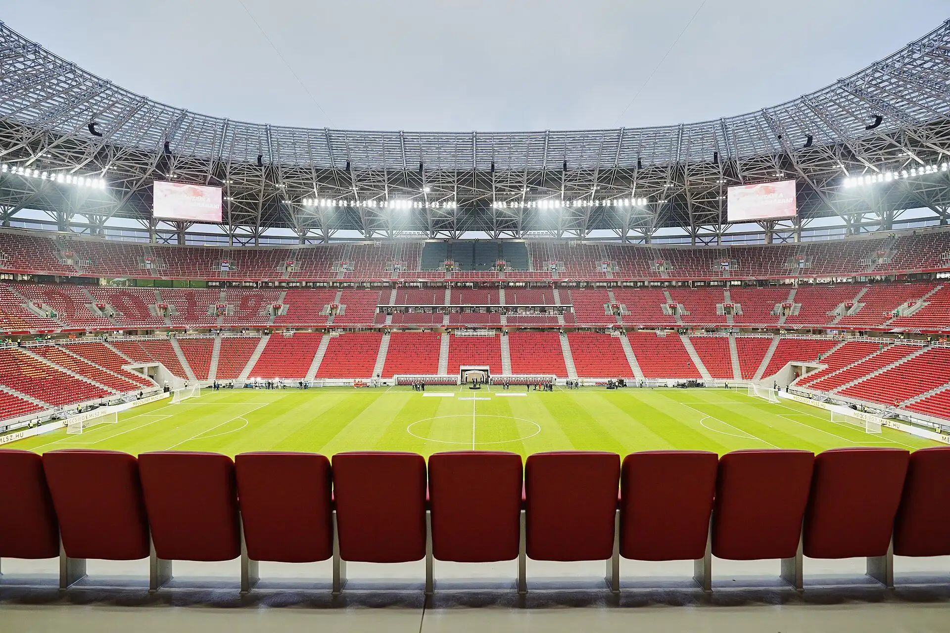 The empty red stands and pitch of Puskás Aréna seen from the lower tier, with the roof structure above
