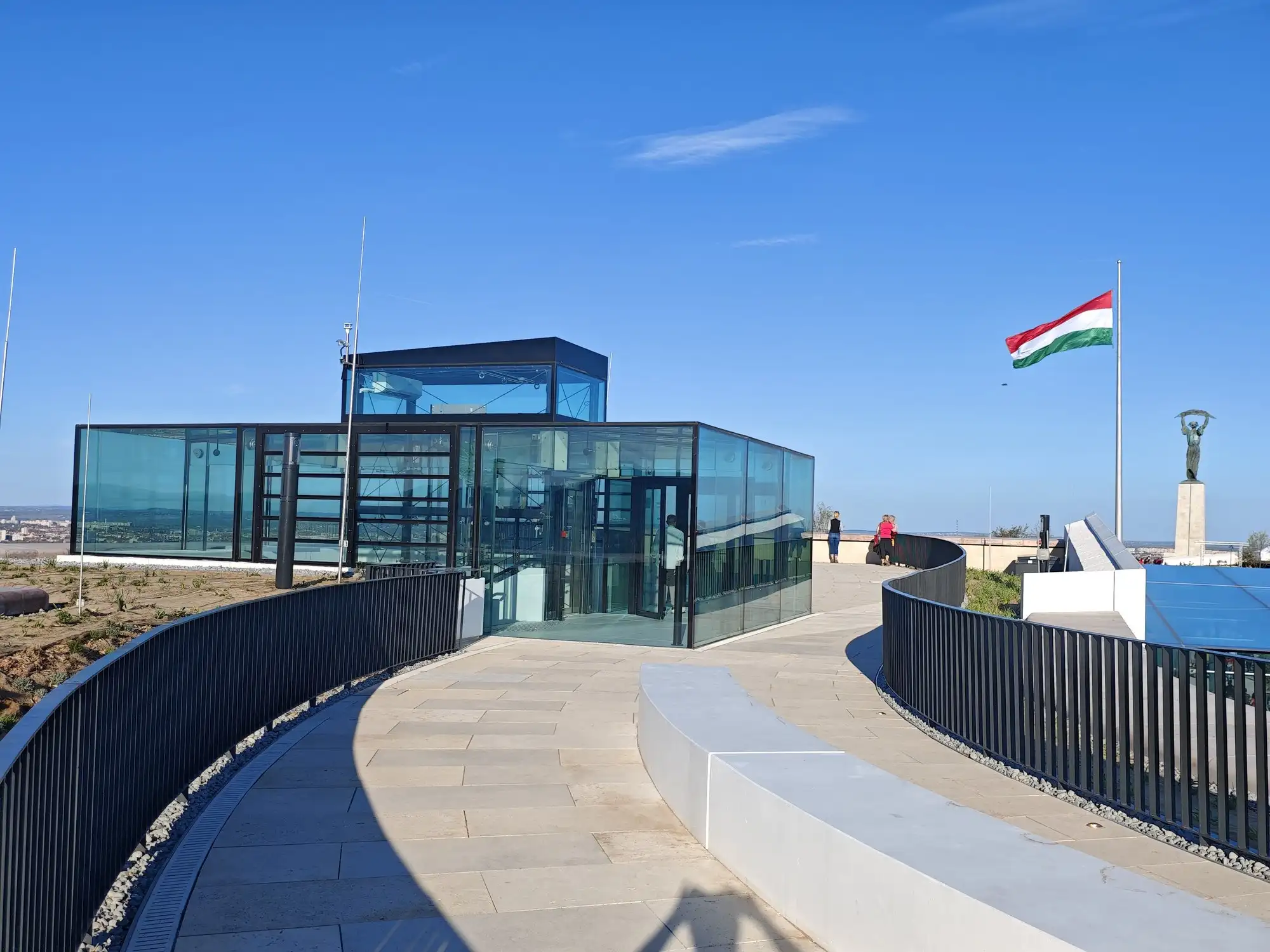 The Roof Garden 360 on top of the Citadella's Western Round Bastion, a curved paved walkway with railings round a glass lift pavilion, the national flag and the Liberty Statue beyond