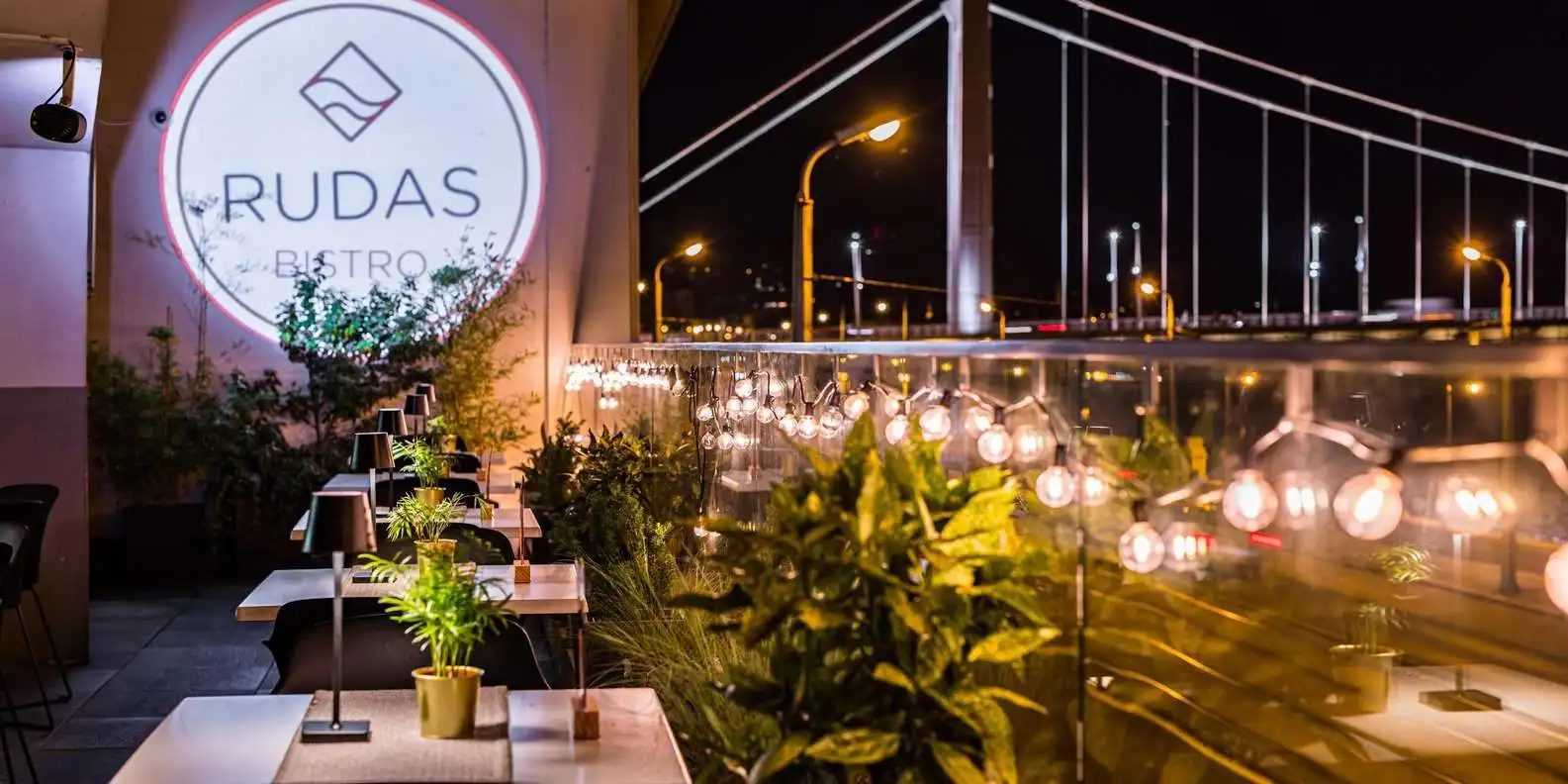 The terrace of Rudas Bistro at night, with tables, plants and string lights, and Elisabeth Bridge lit up behind