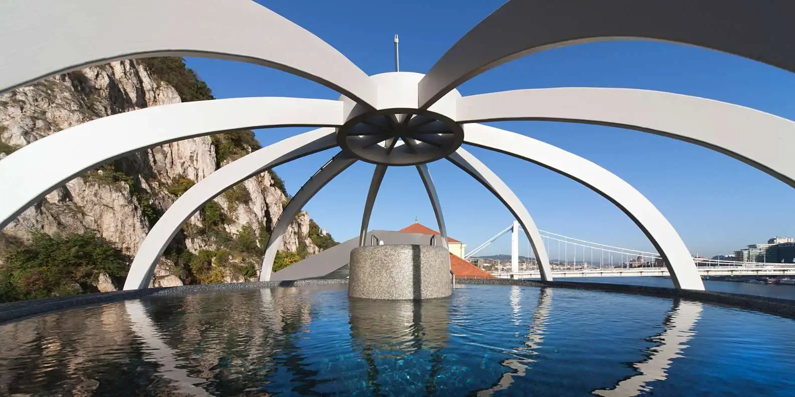 The rooftop panorama pool at Rudas Thermal Bath under a white ribbed canopy, with Elisabeth Bridge and Gellért Hill's cliffs behind