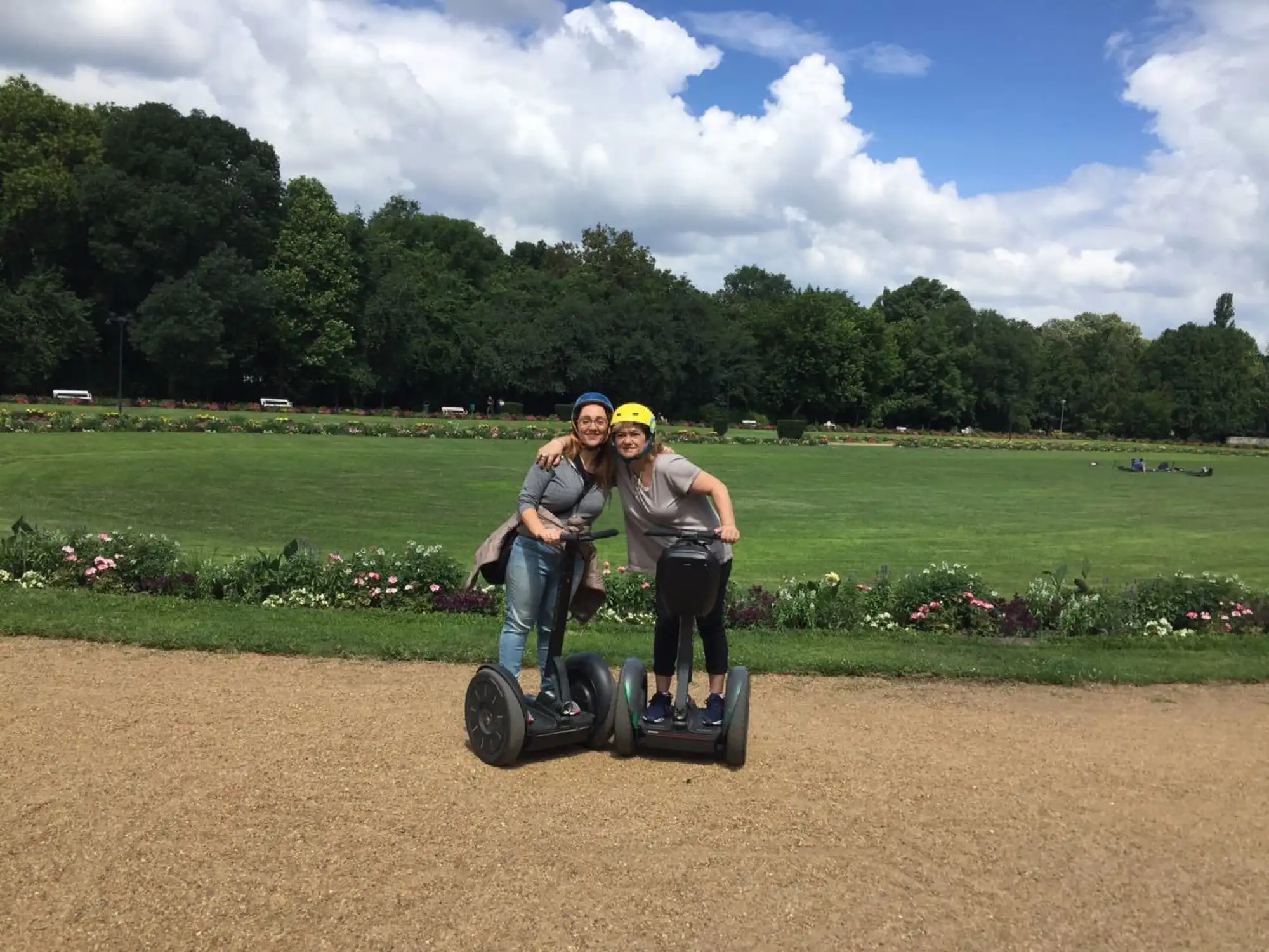 Two riders in helmets on Segways on a gravel path on Margaret Island, with a wide lawn, flower beds and a line of trees behind them