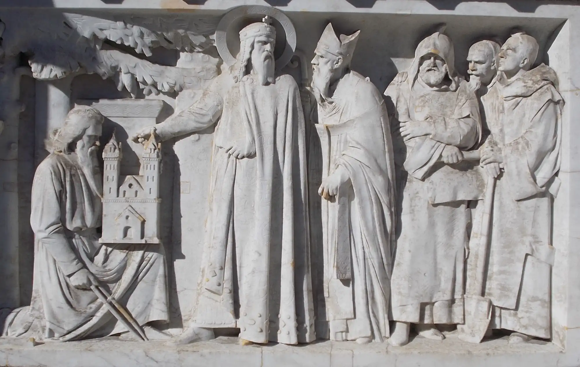 A white stone relief on the plinth of St Stephen's statue, showing a bearded master builder kneeling before the king with a model of a church in his hands