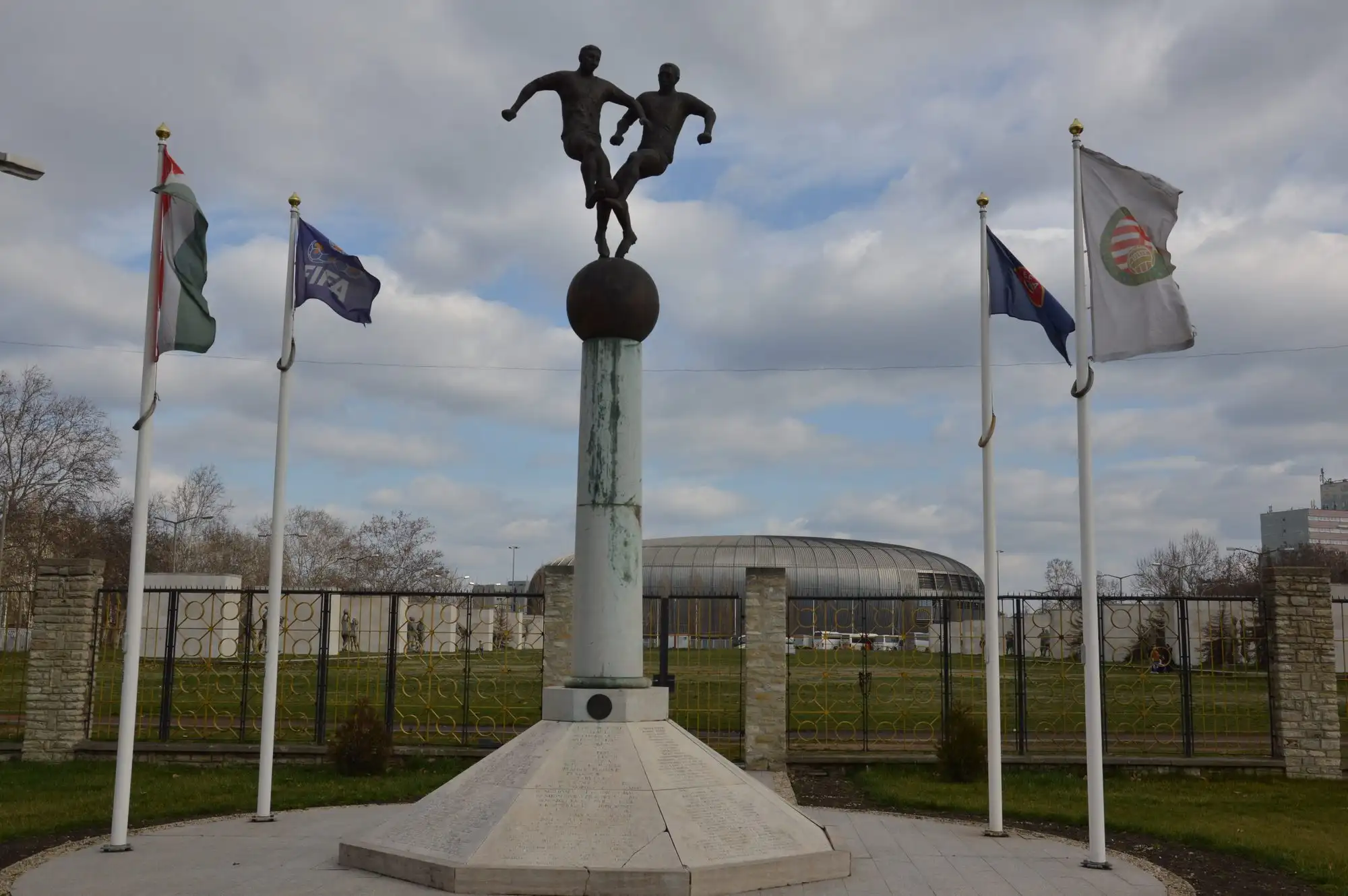 The Statue of Hungarian Football beside Puskás Aréna: two bronze players on a globe on a tall plinth, with flags flying either side
