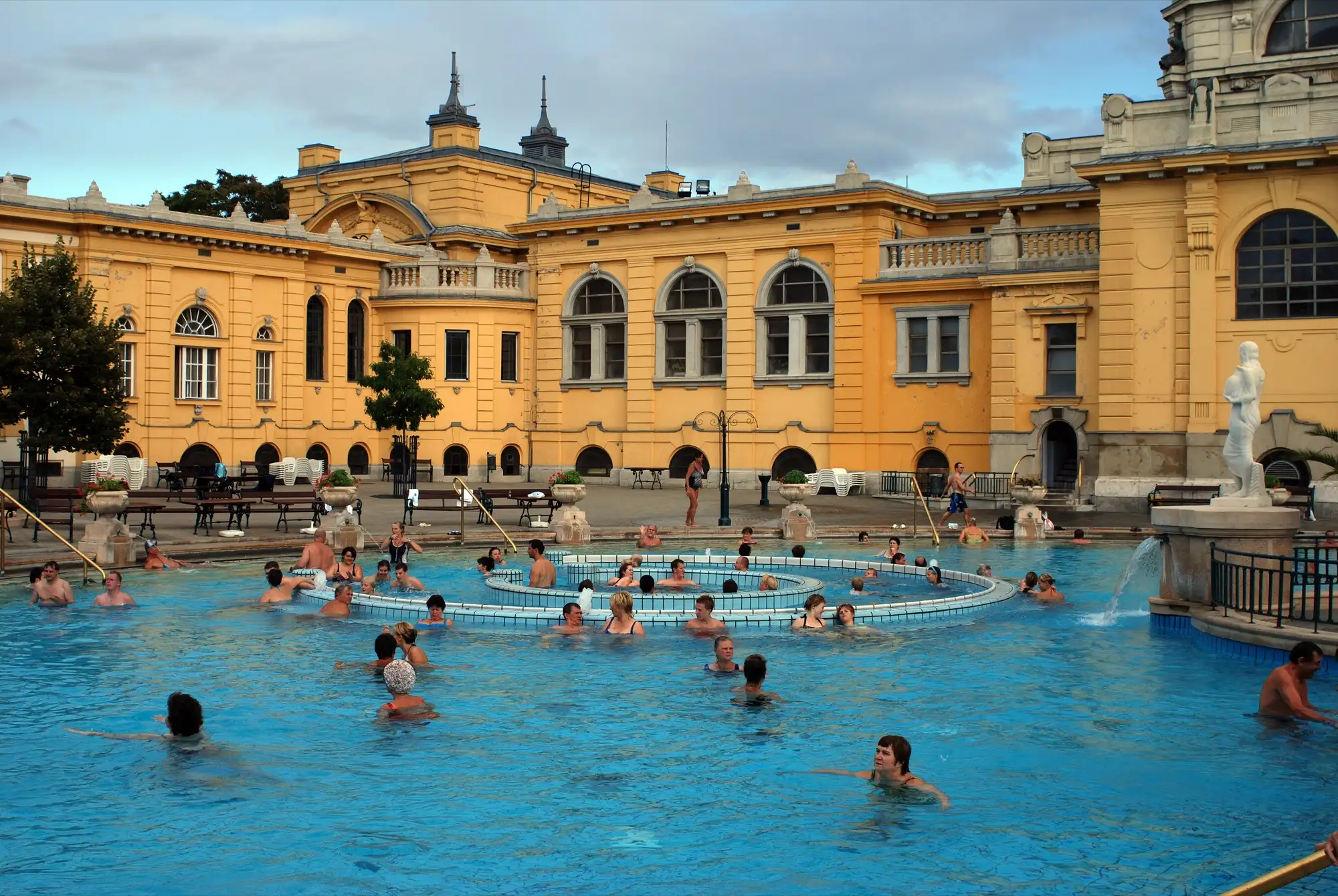 Bathers in the outdoor leisure pool at Széchenyi Thermal Bath, some riding the circular whirling corridor in the middle, with the yellow bath buildings behind