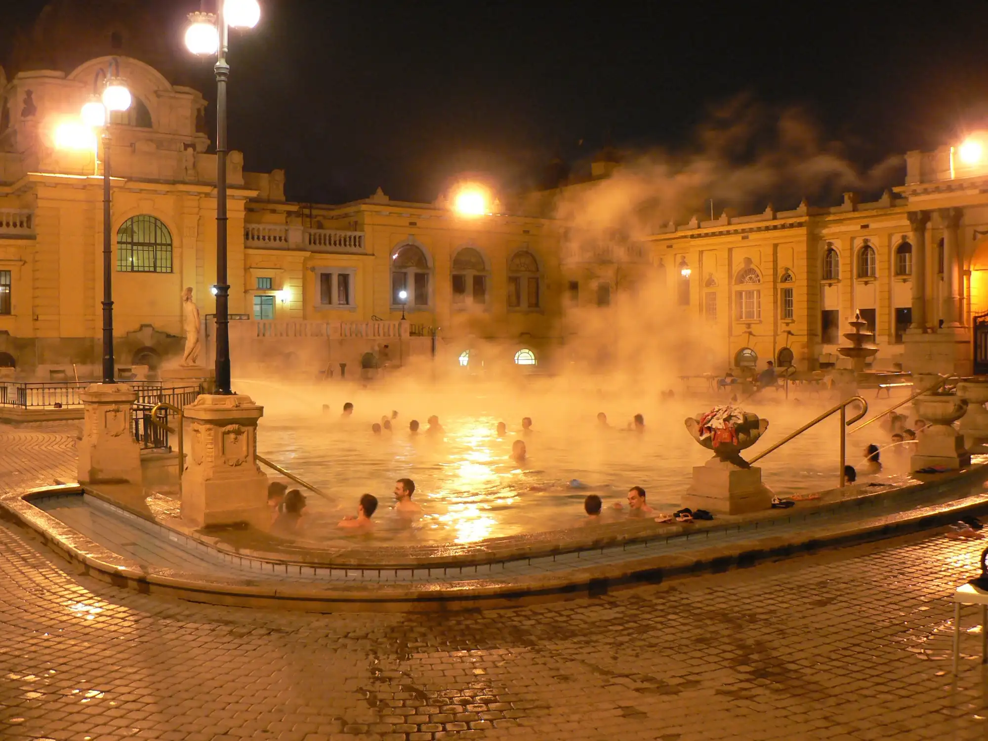 Steam rising off the hot outdoor pool at Széchenyi Thermal Bath on a winter night, with bathers in the floodlit water and the yellow bath buildings behind