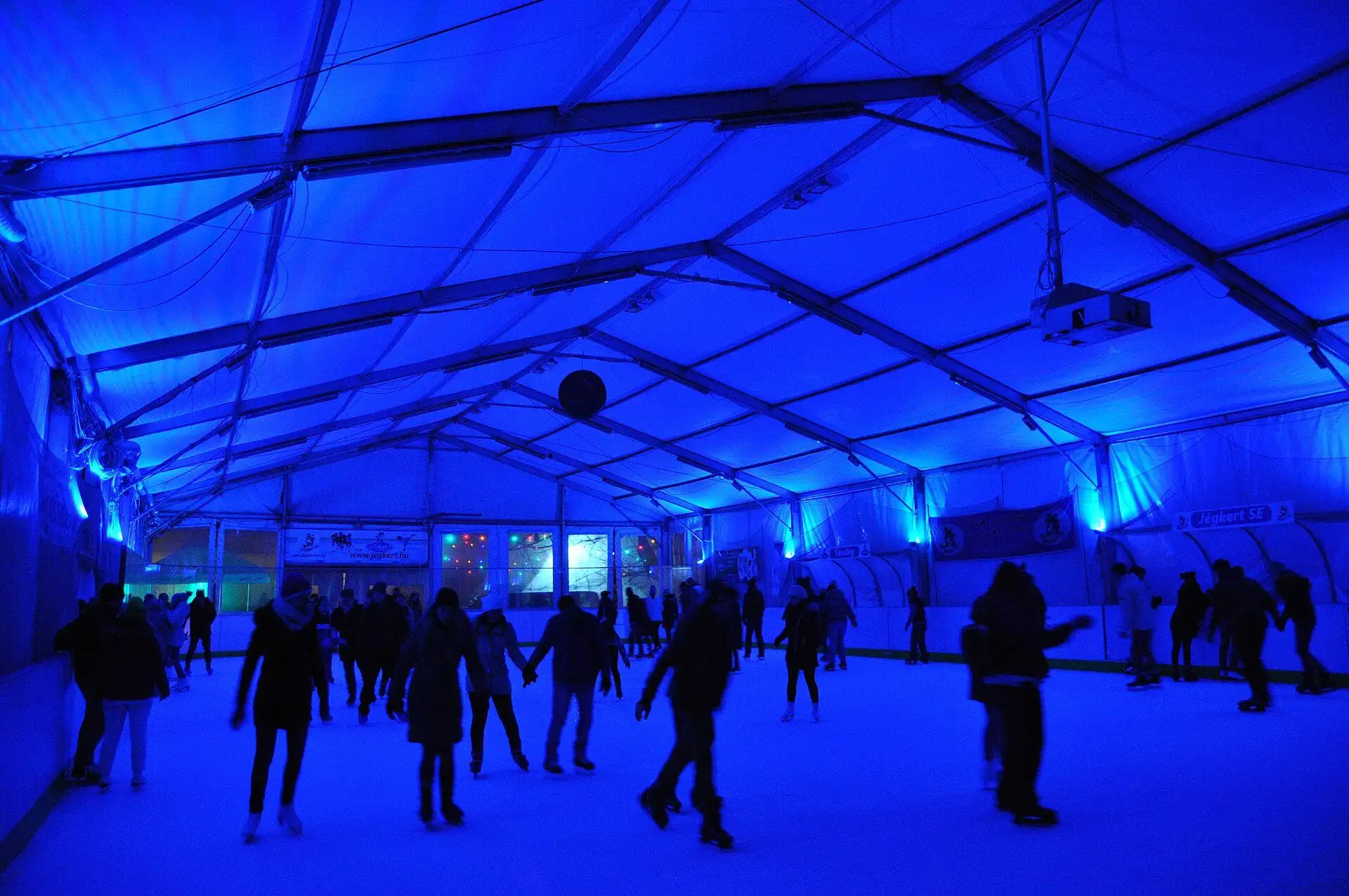 Skaters under the blue-lit roof of the covered Jégkert ice rink on Széna tér in Budapest at night