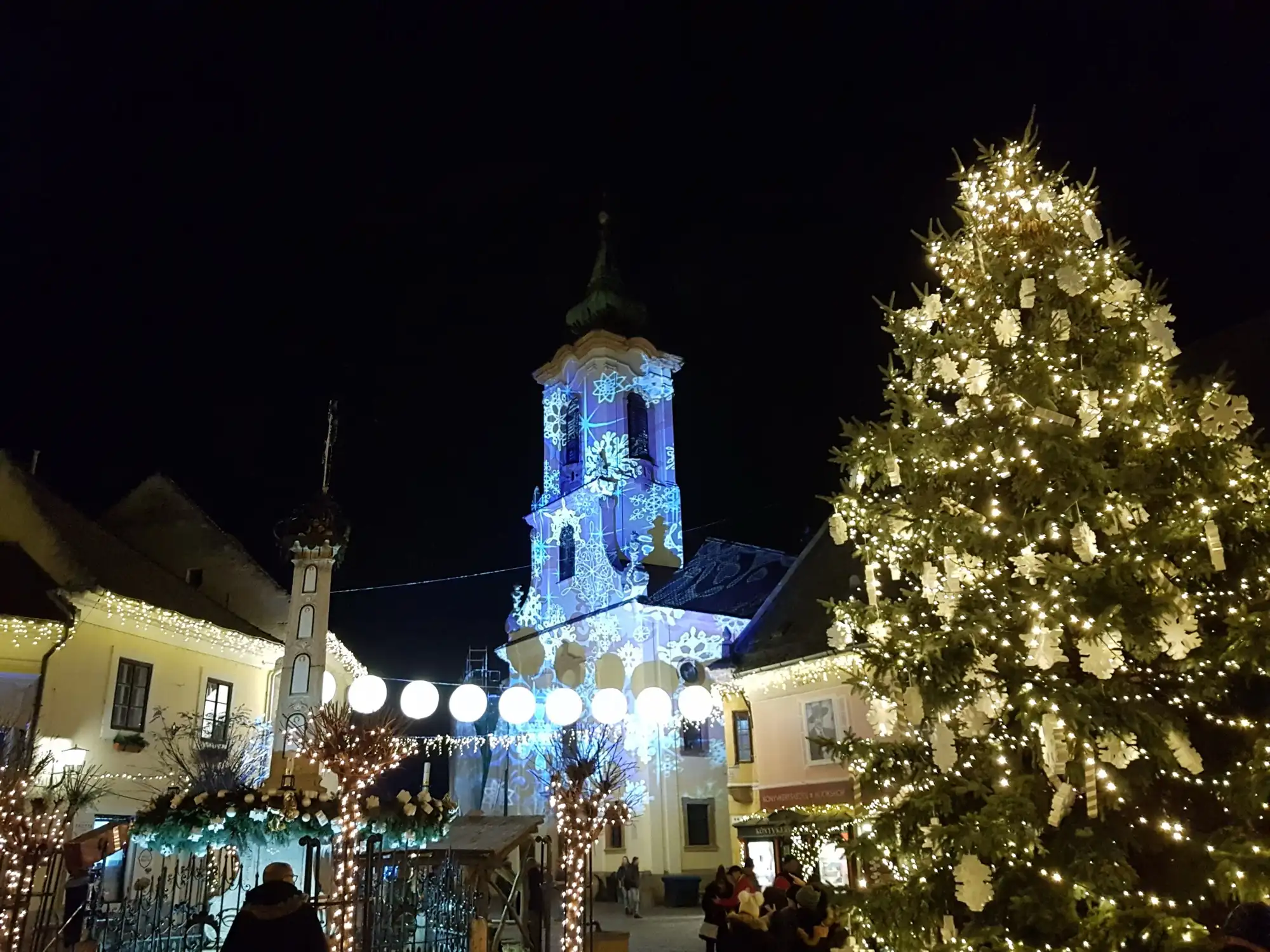 The lit Christmas tree on Fő tér in Szentendre at night, with a church tower lit by projected snowflakes behind it