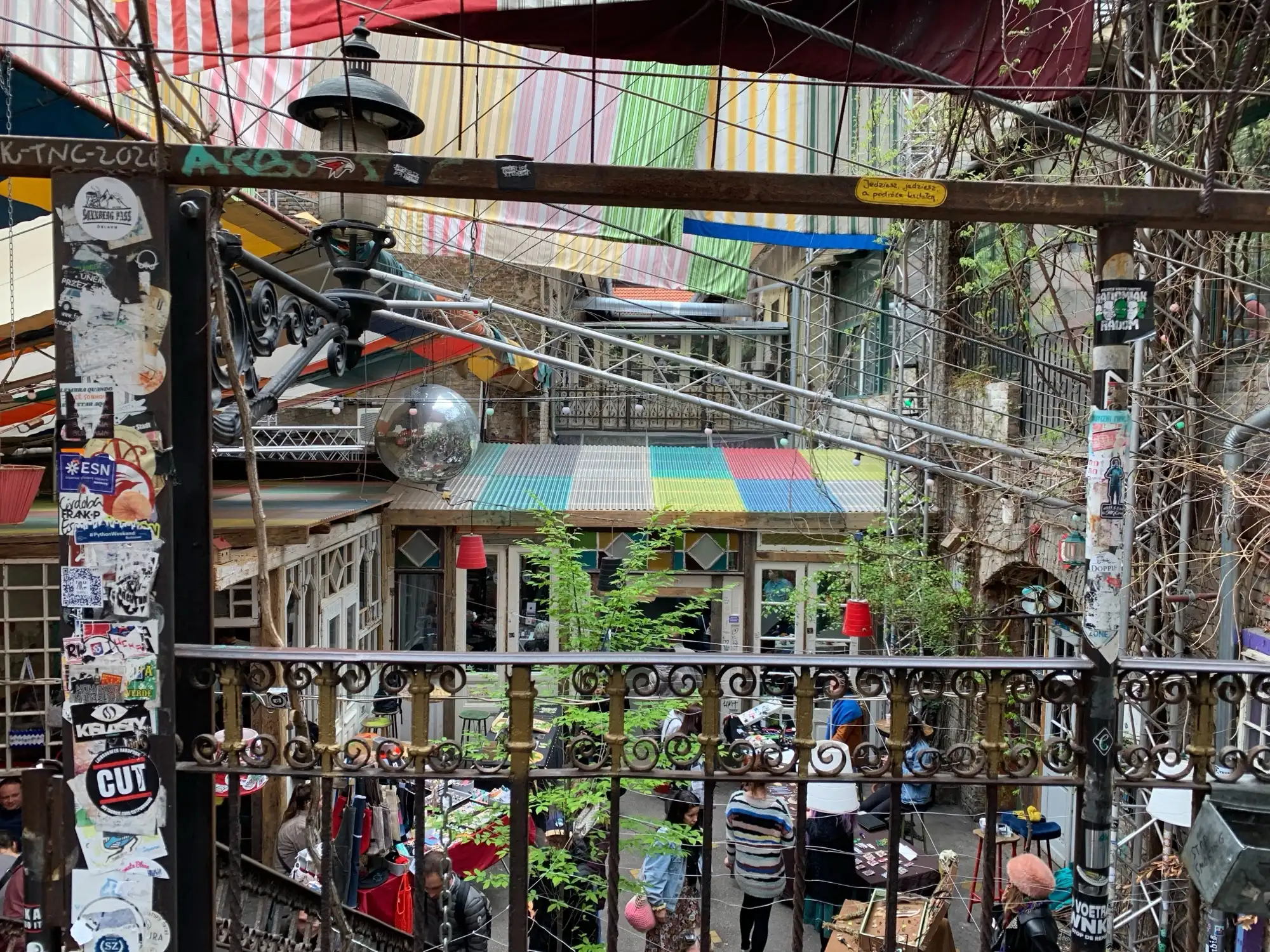 Looking down into the courtyard of Szimpla Kert in daylight, past a sticker-covered post and an iron railing, to striped awnings, a mirror ball and market stalls below
