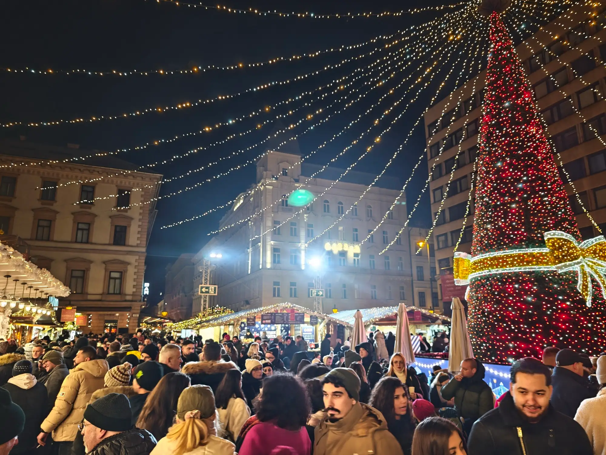 The tall Christmas tree at Advent Bazilika wrapped in lights, with strings of lights fanning out from its top over a crowd on Szent István tér at night