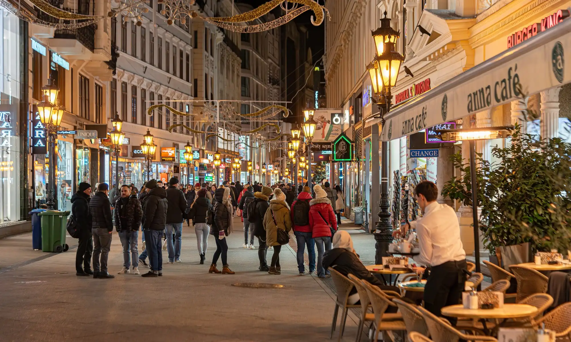 Váci utca at night in late November, with gold garlands of lights strung across the pedestrian street, lit shop windows and people walking