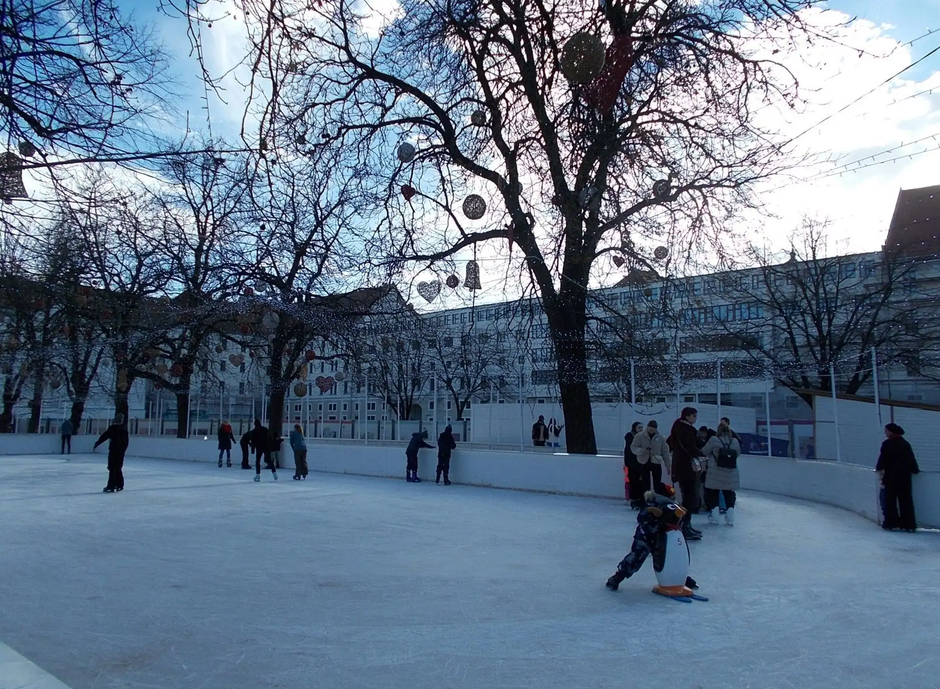 Skaters on the outdoor rink in Városháza Park on a sunny winter afternoon, with Christmas baubles hanging in the bare trees and the Budapest City Hall behind