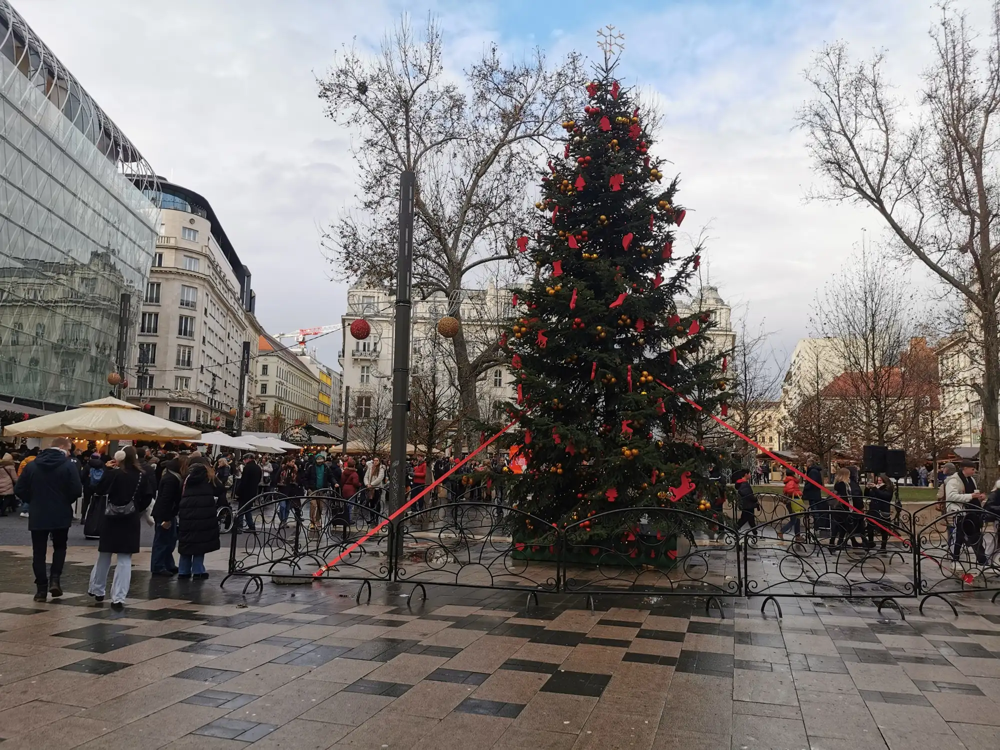 The Christmas tree on Vörösmarty tér decorated in red and gold by day, with shoppers among the market stalls behind it