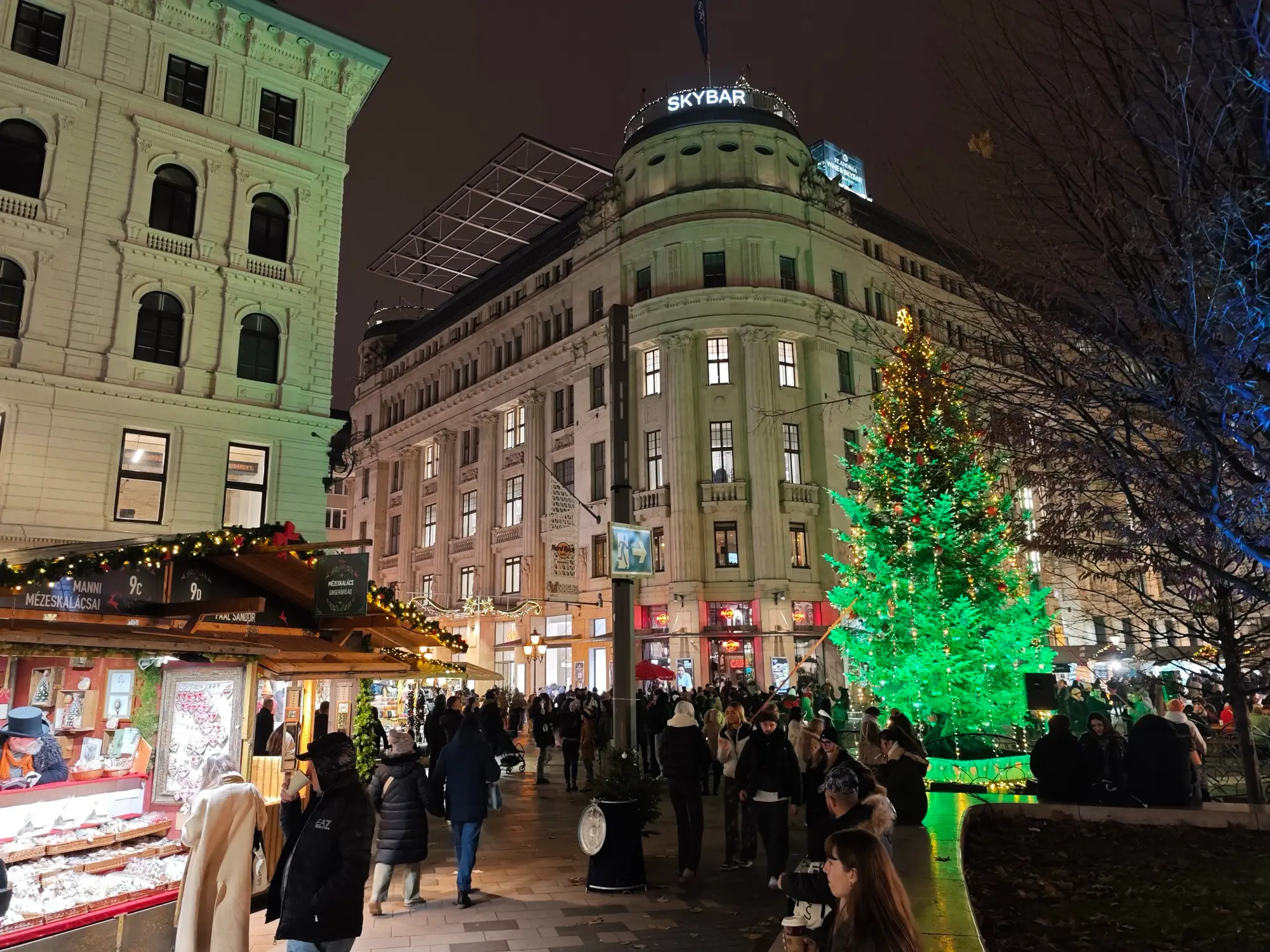 The Christmas tree lit green on Vörösmarty tér at night, with market stalls and crowds in front of the surrounding buildings