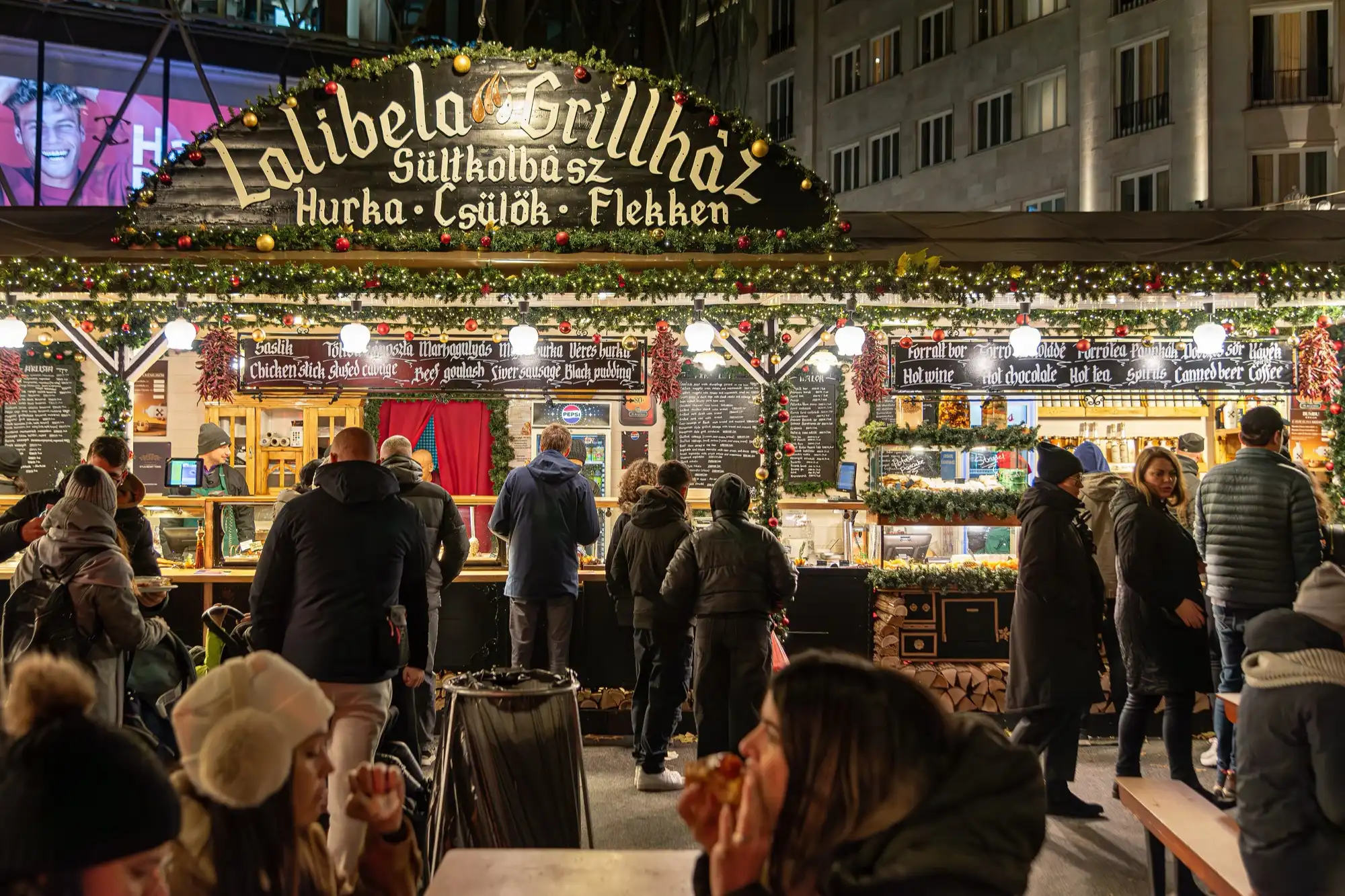 A grill stall decorated with Christmas garlands at the Vörösmarty tér market, with customers queuing at the counter