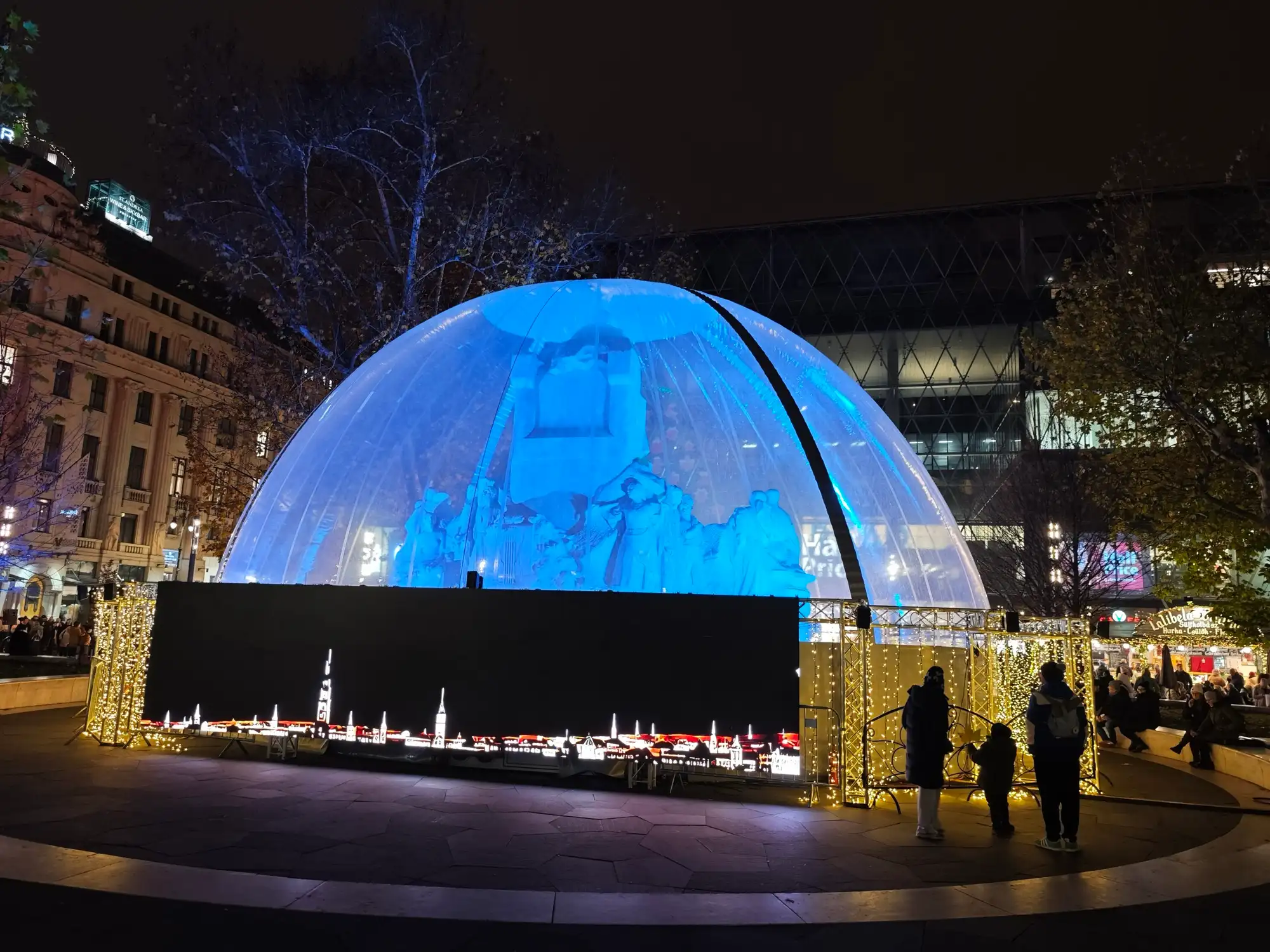 The statue in the middle of Vörösmarty tér inside a transparent dome lit blue at night, with an LED wall of the Budapest skyline in front