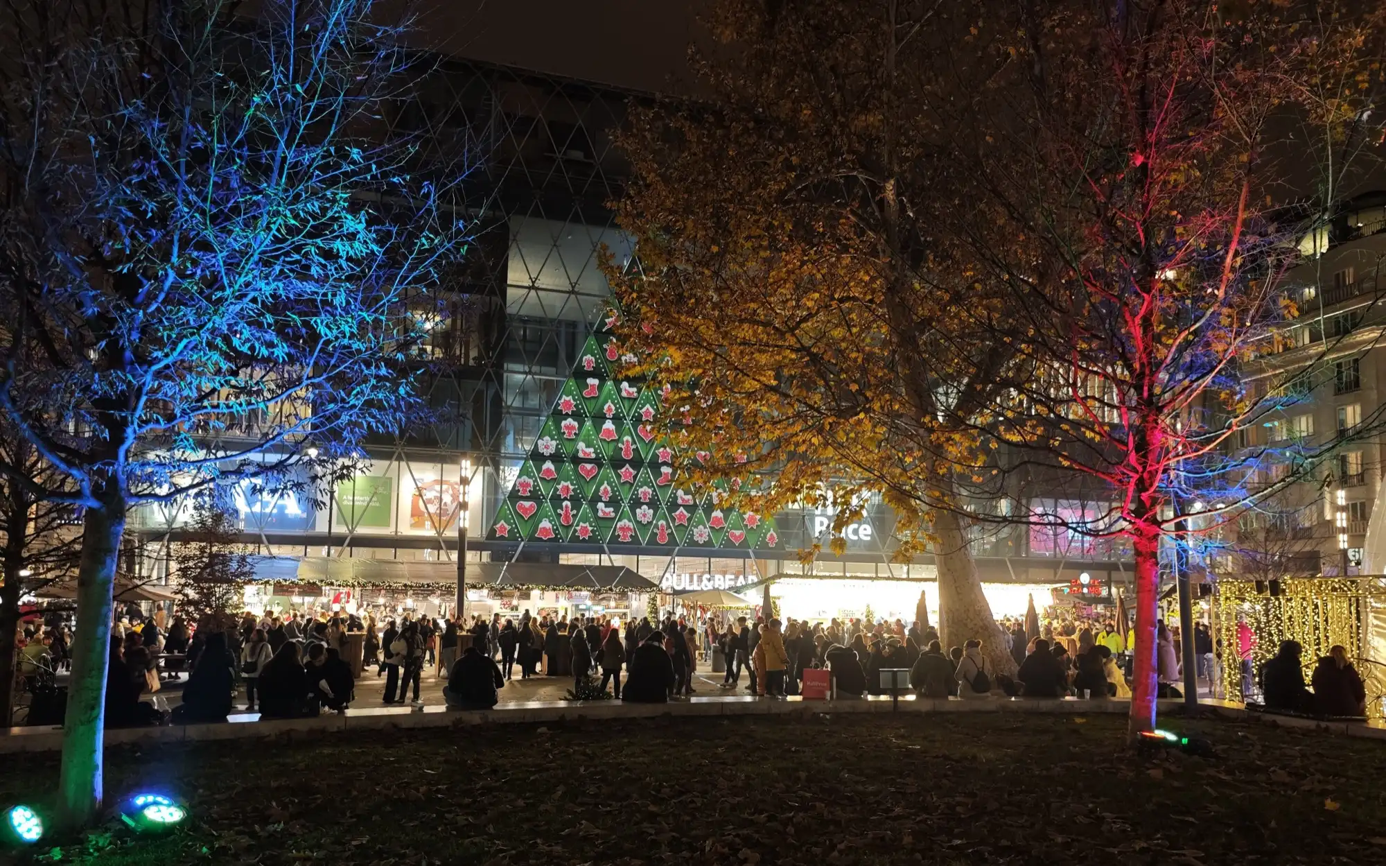 Trees on Vörösmarty tér uplit in blue and red at night, with a Christmas tree of lights on the glass front of a building and the market stalls lit below