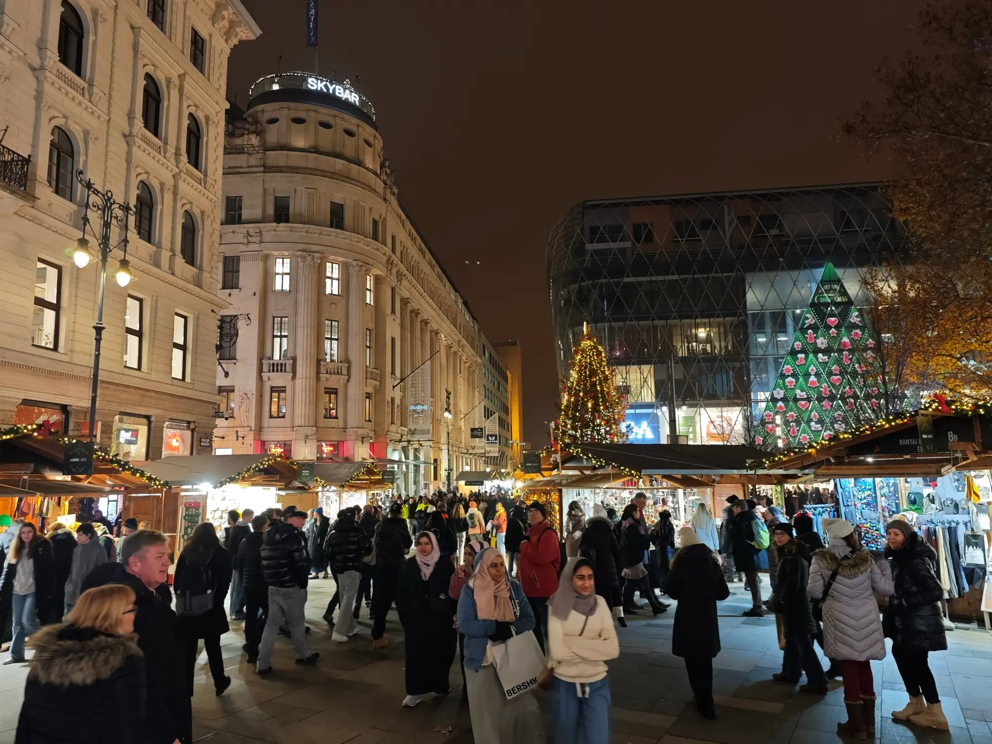 Crowds walking between the lit stalls of the Vörösmarty tér Christmas fair at night