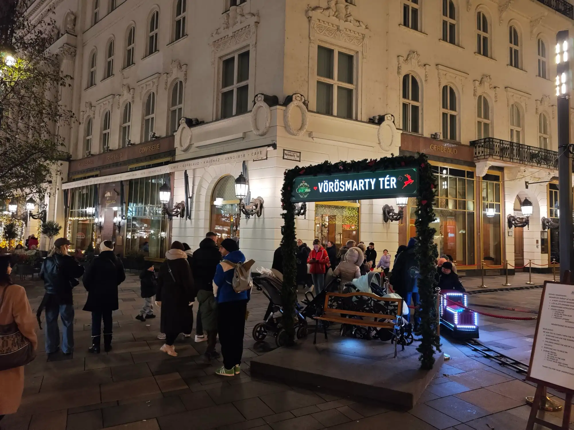 The lit Vörösmarty tér stop of the children's train at night, with the little train and families waiting in front of the Gerbeaud building