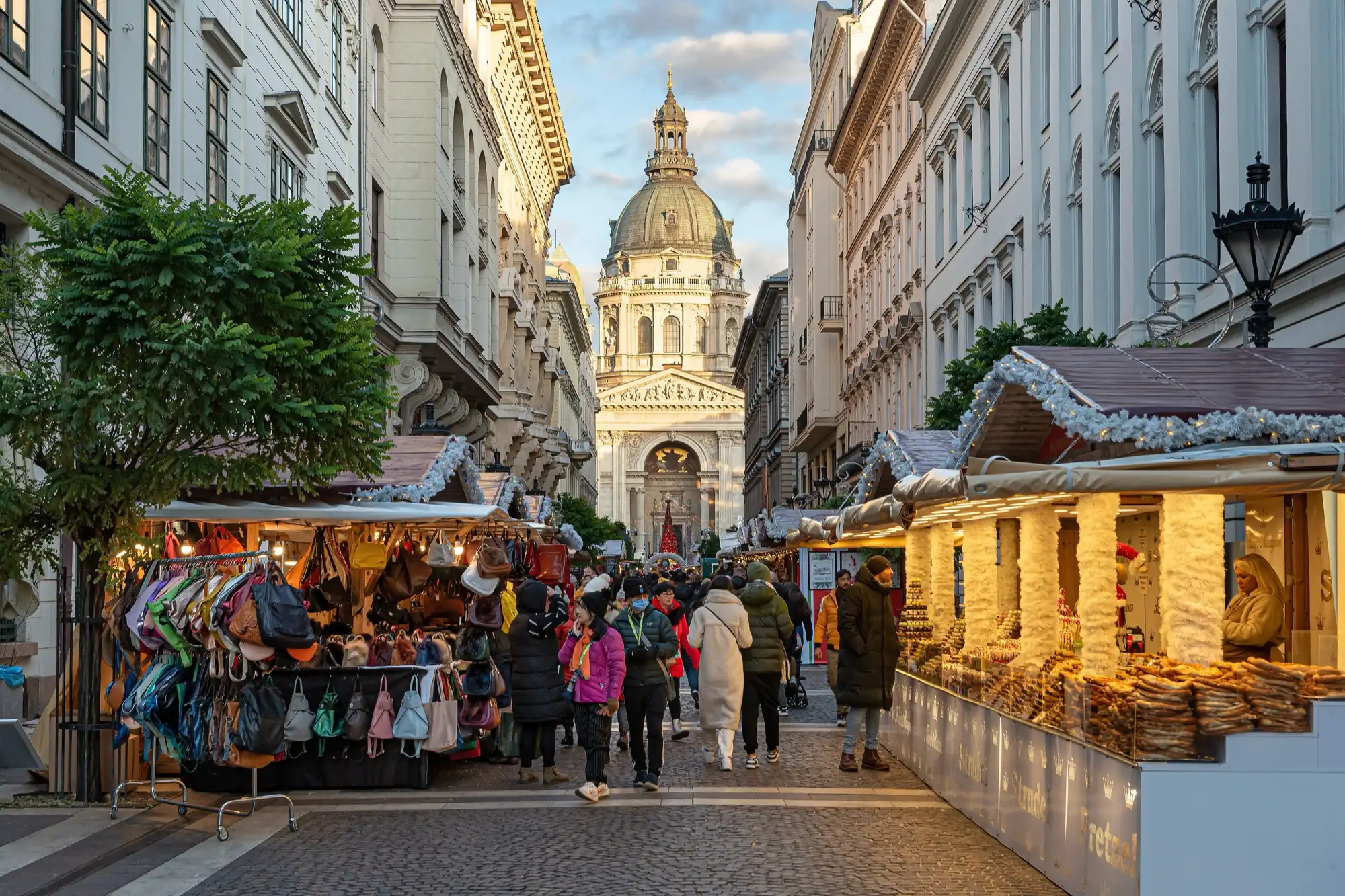 Christmas market stalls selling bags and pastries along Zrínyi utca in late-afternoon light, with the dome and front of St Stephen's Basilica at the end of the street