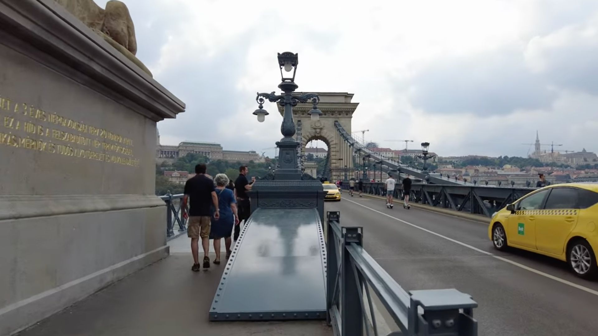 People walking on the Széchenyi Chain Bridge in Budapest with a view of the lion statues and cityscape in the background.