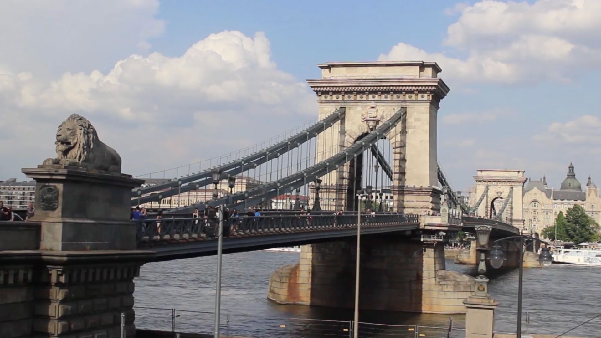 The Széchenyi Chain Bridge in Budapest with its iconic lion statues and architectural details, set against a partly cloudy sky.