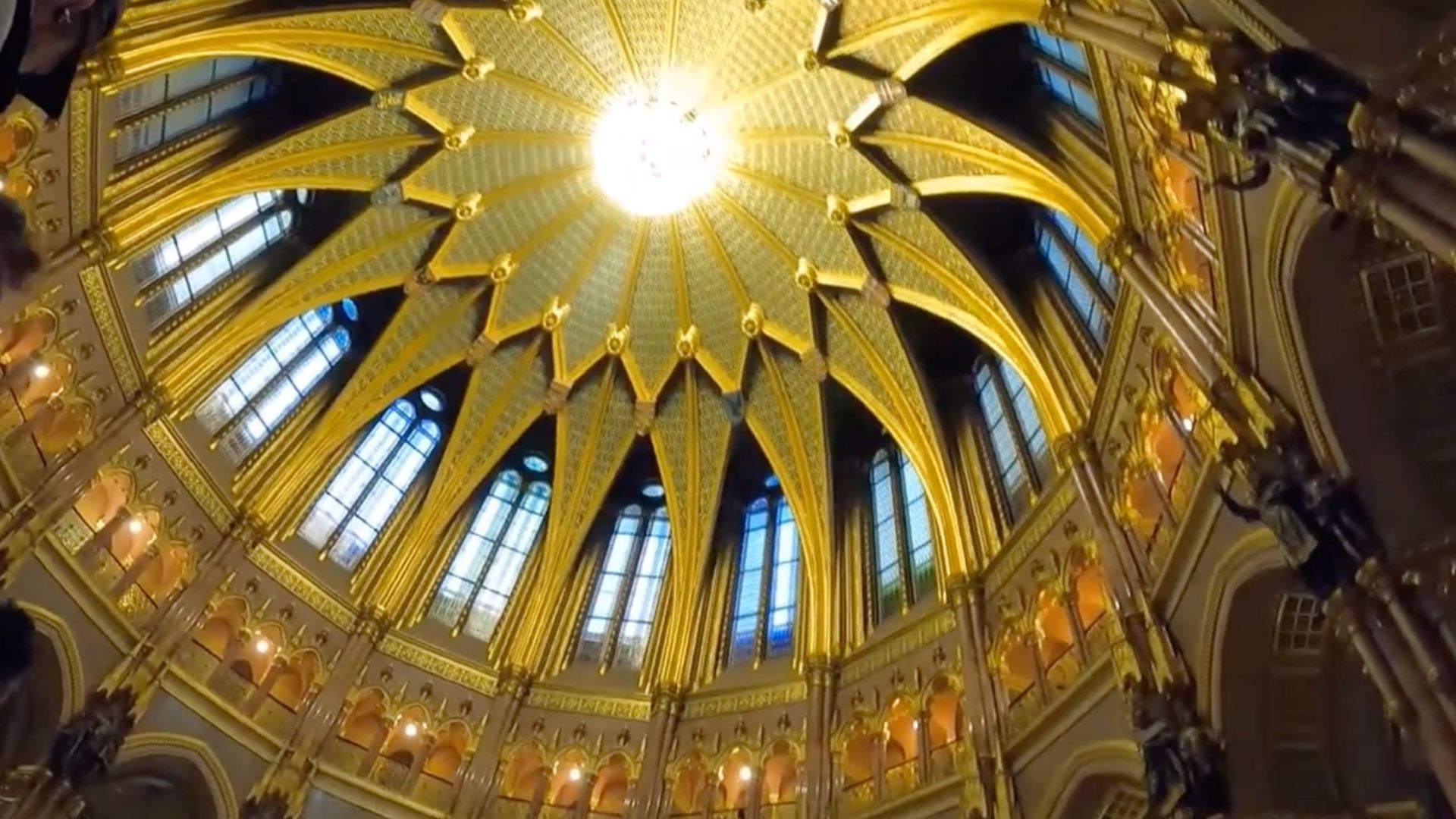 Hungarian Parliament Building interior view of the ornate dome ceiling with golden arches and intricate patterns illuminated by a central light fixture.