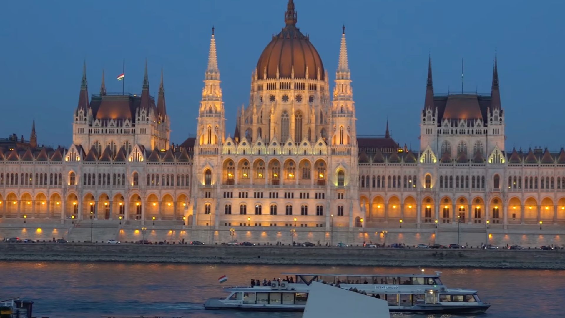 Evening view of the Hungarian Parliament Building with illuminated gothic architecture, reflecting in the Danube River, with boats passing in the foreground.