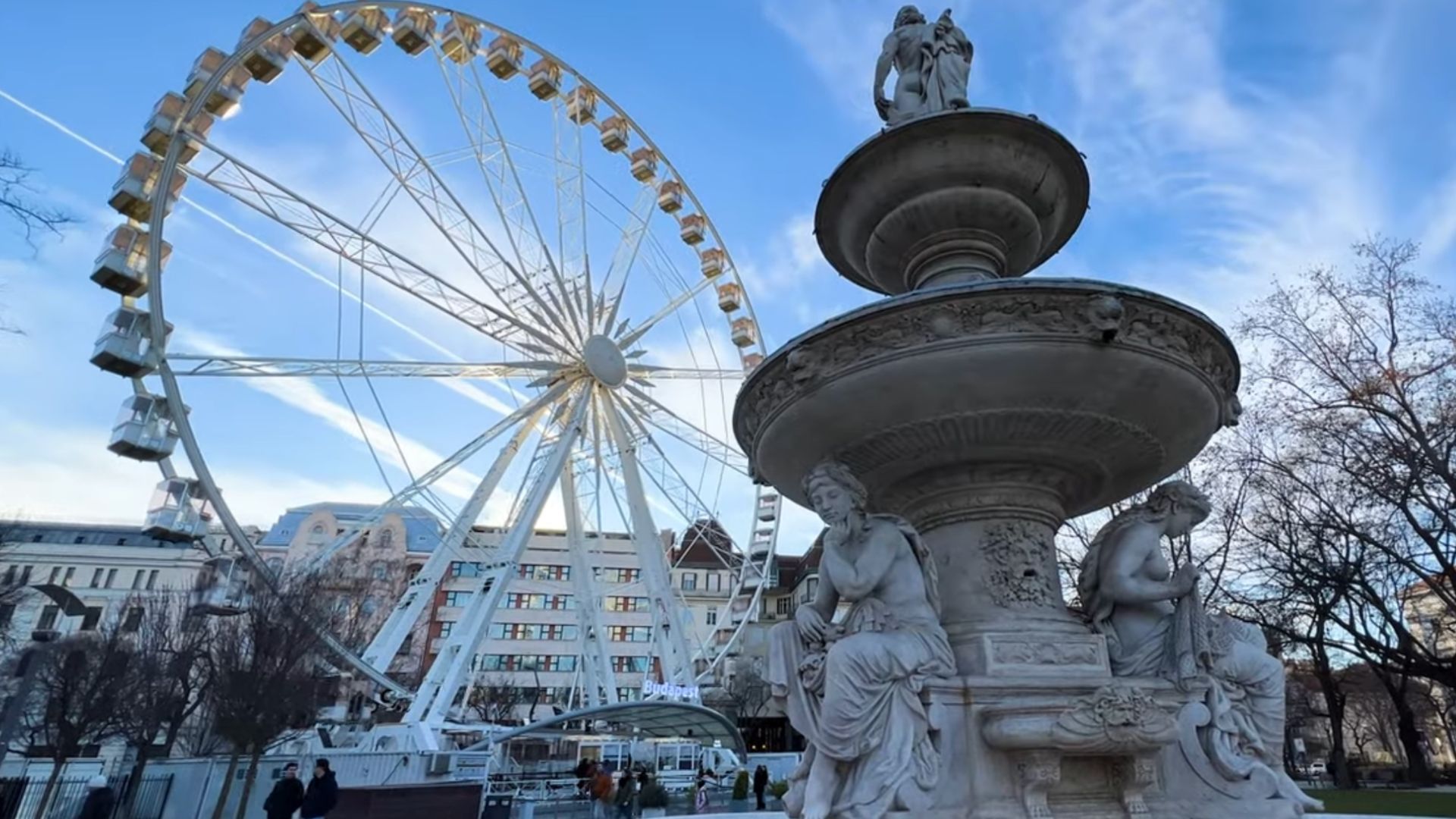 Budapest Eye ferris wheel behind a decorative fountain with statues, set against a clear blue sky.