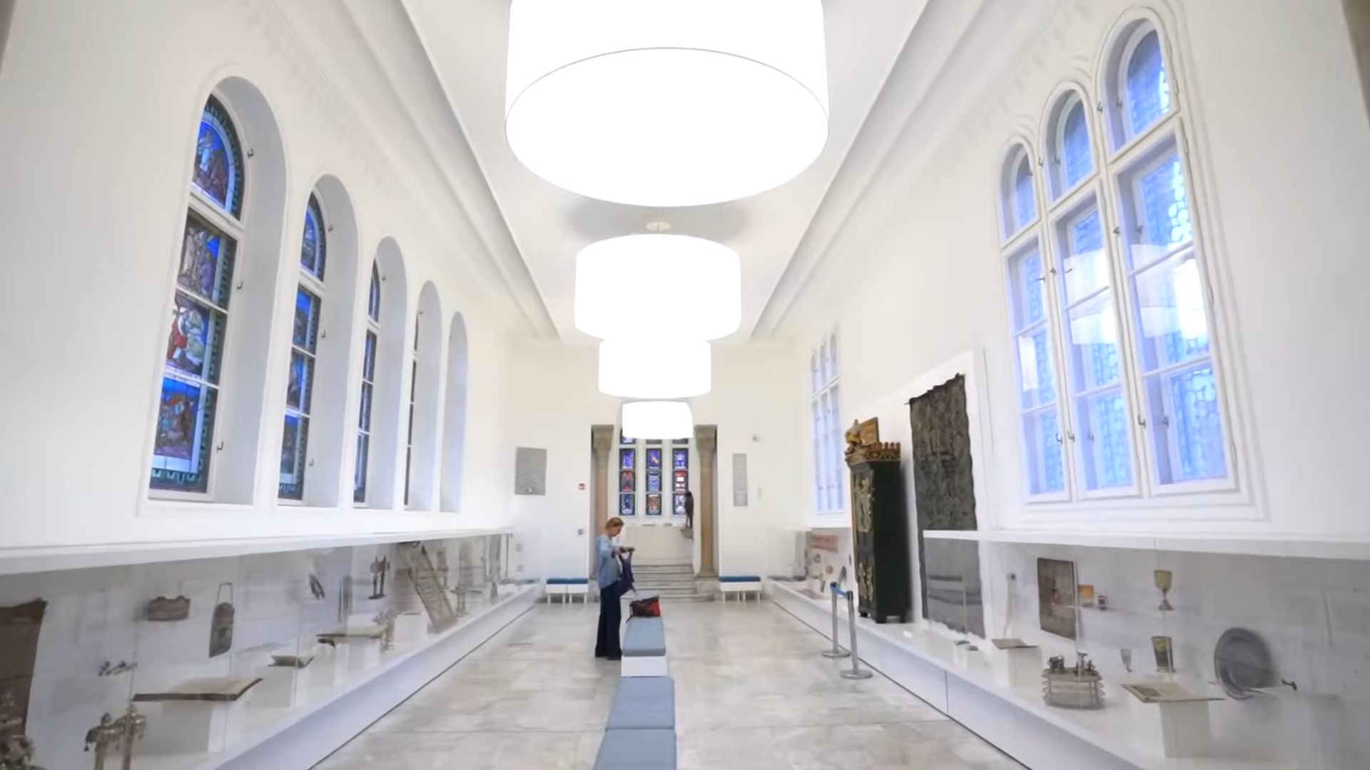 A white-walled museum hall inside the Dohány Street Synagogue in Budapest, featuring stained glass windows, exhibits along the walls, and large overhead lighting.