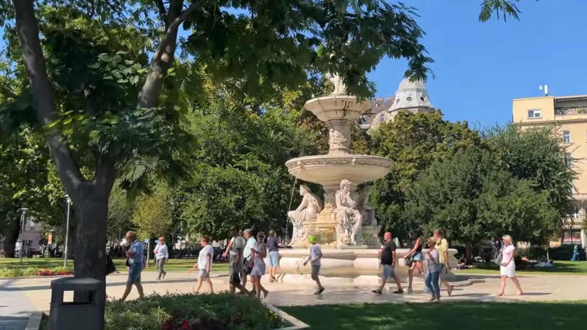 People walking around a decorative fountain in Erzsebet Square in Budapest, with lush green trees and buildings in the background.