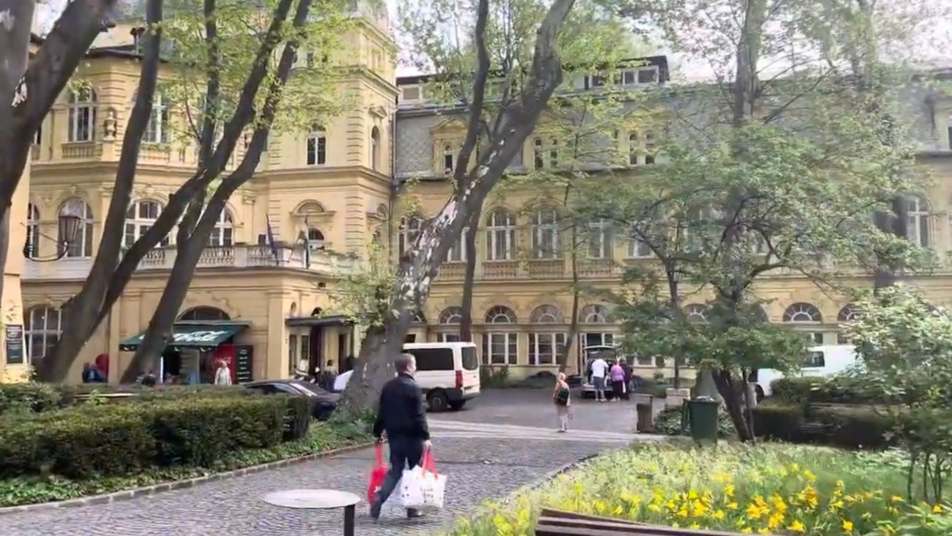 View of Lukács Thermal Bath's garden with green trees, yellow flowers, and the historic bath building in the background.